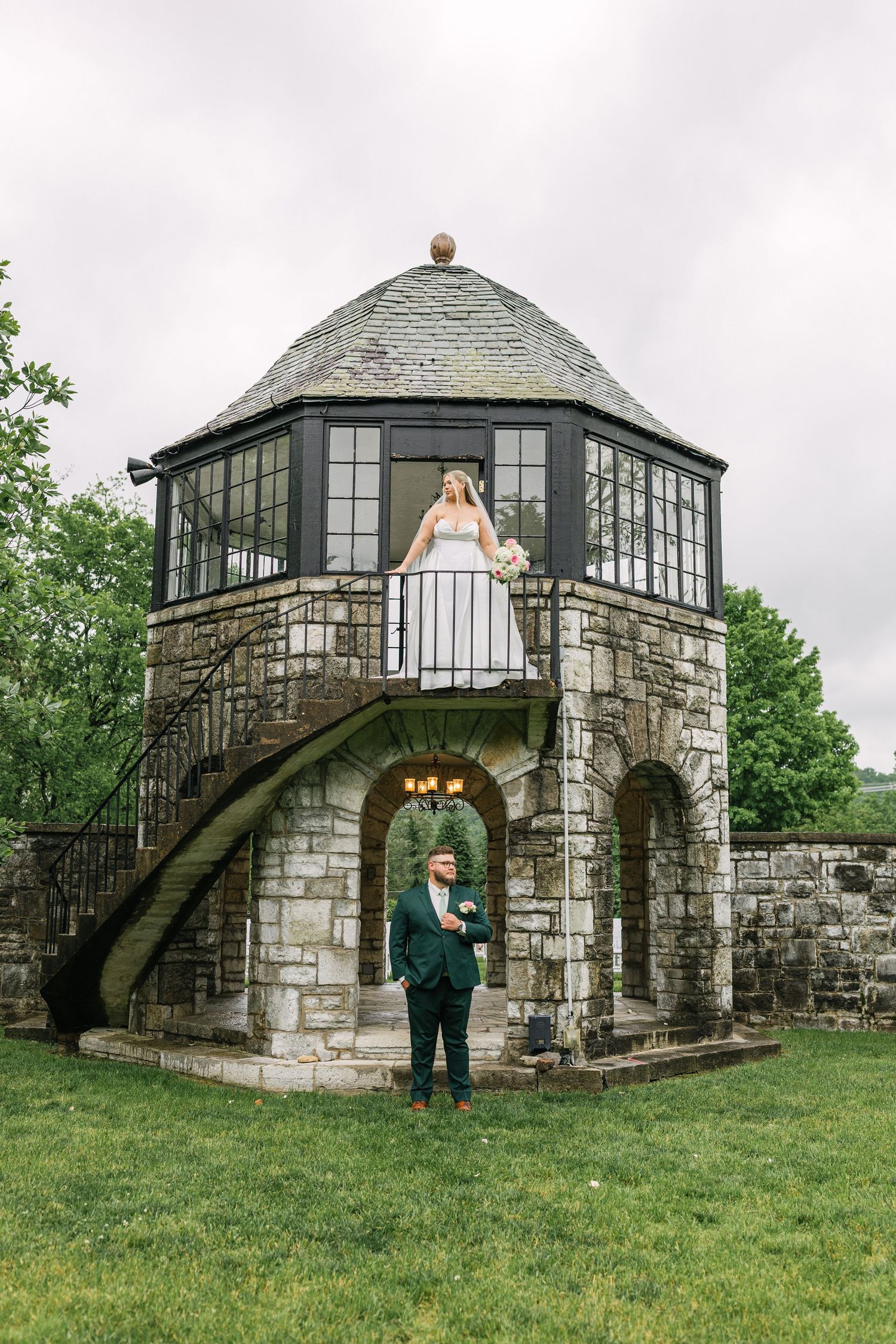 Bride on balcony of stone tower with groom standing below on green lawn at elegant outdoor wedding venue.