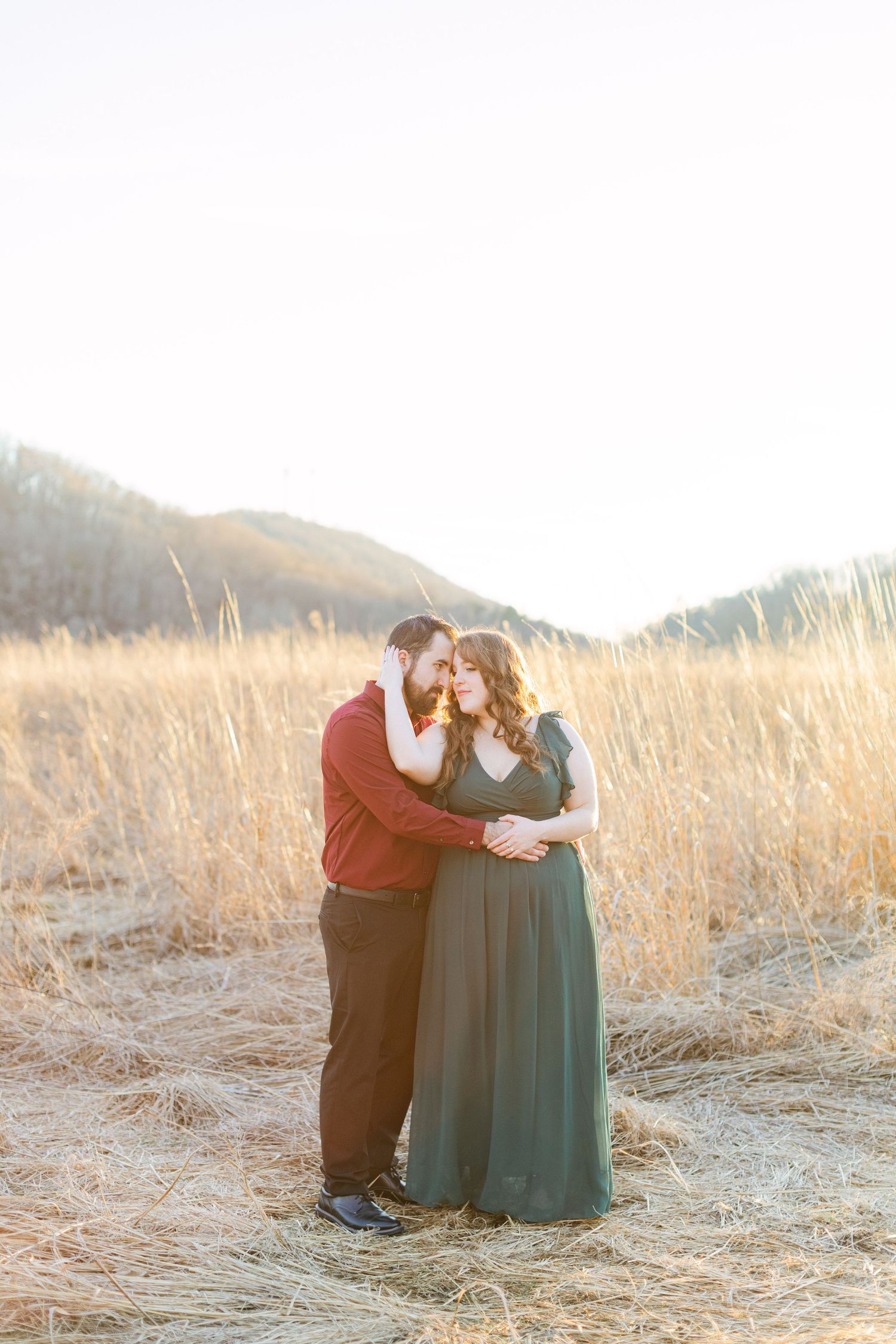 Couple embracing in golden field at sunset, woman in green gown, man in burgundy shirt, mountains in background.