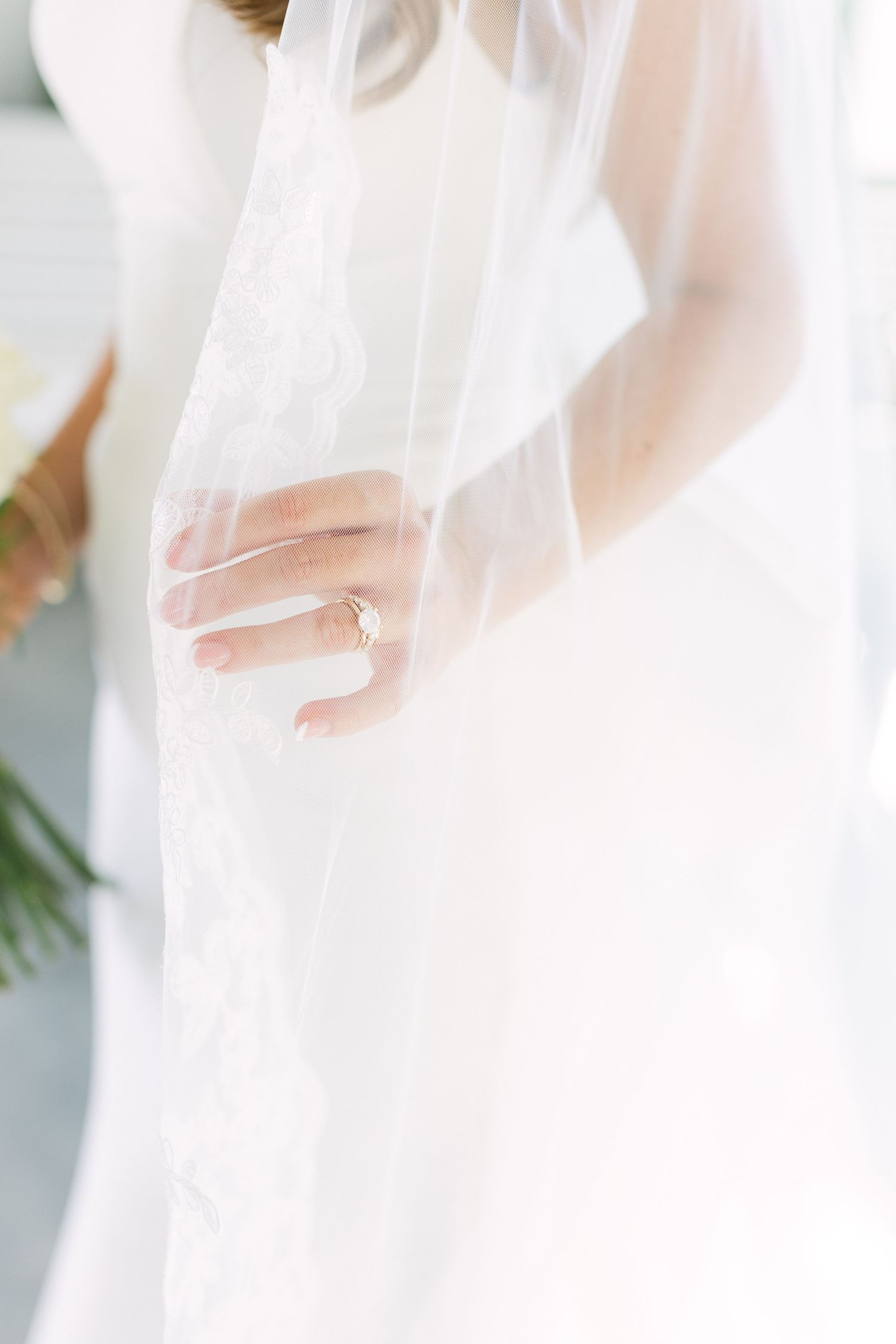 Bride in white wedding gown with lace veil, showing delicate engagement ring on her hand while holding a bouquet.