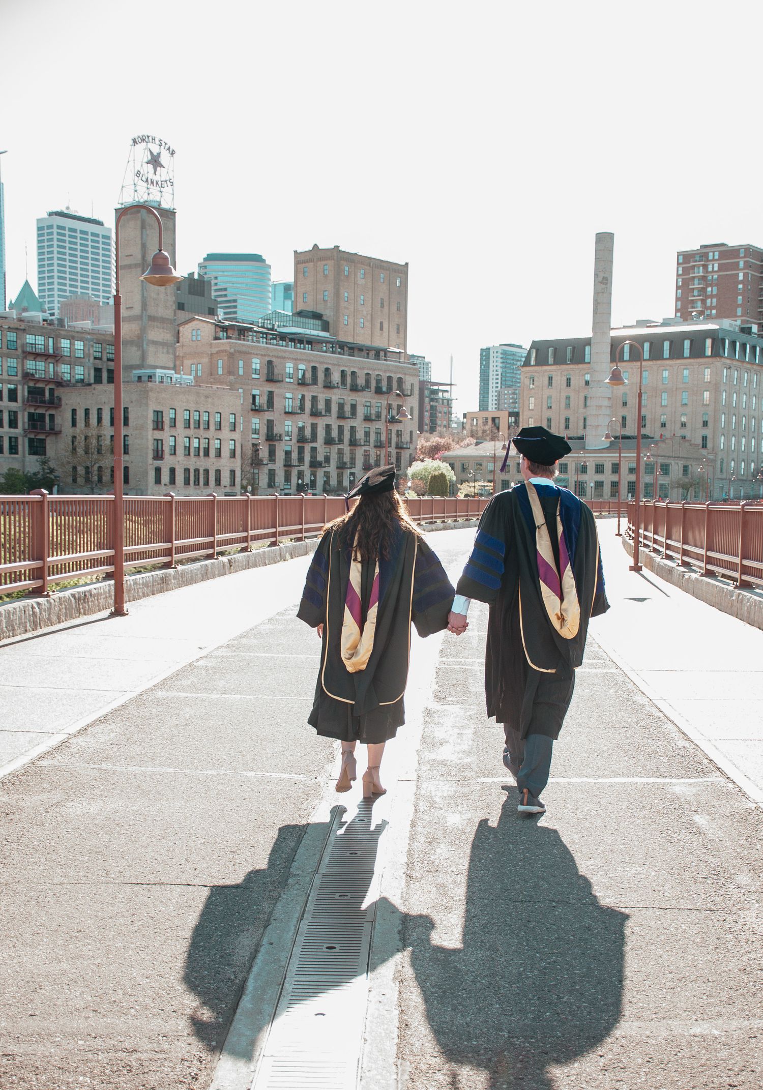 Two graduates walking on bridge wearing black graduation gowns with city skyline in background.