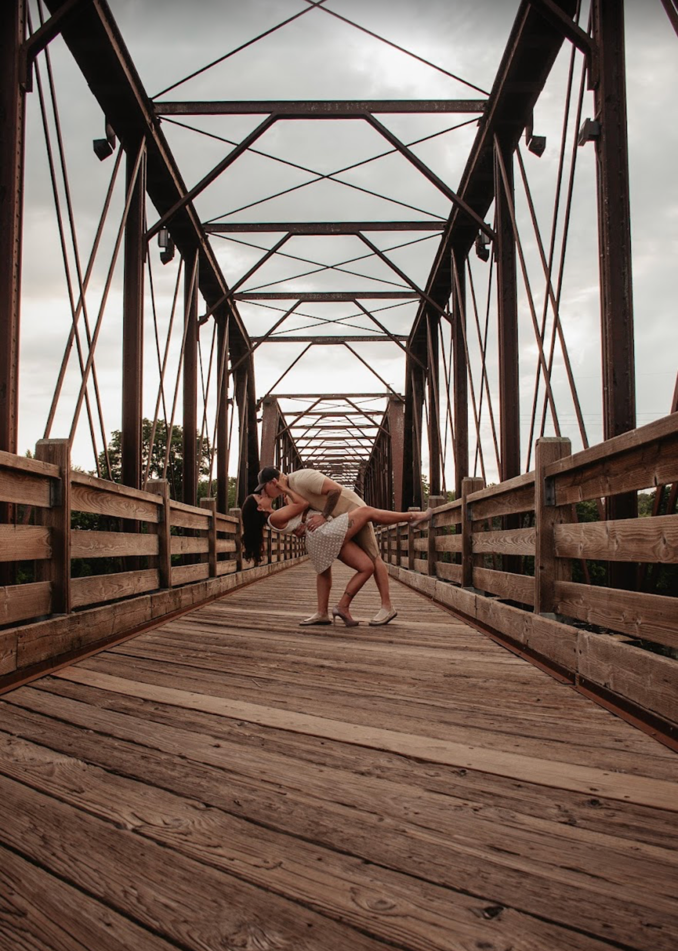 Couple sharing a romantic dip kiss on a rustic wooden bridge with iron truss framework under cloudy skies.