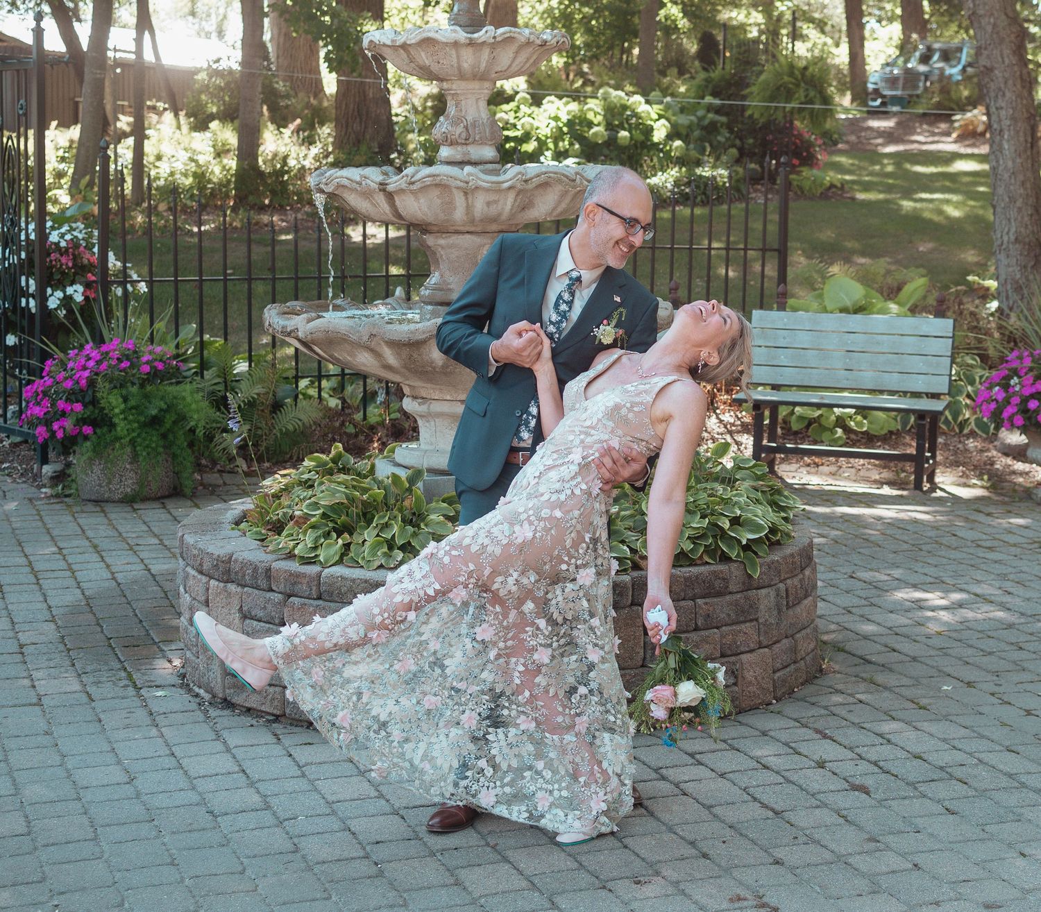 A romantic dance dip pose in front of a garden fountain with purple flowers and brick landscaping.