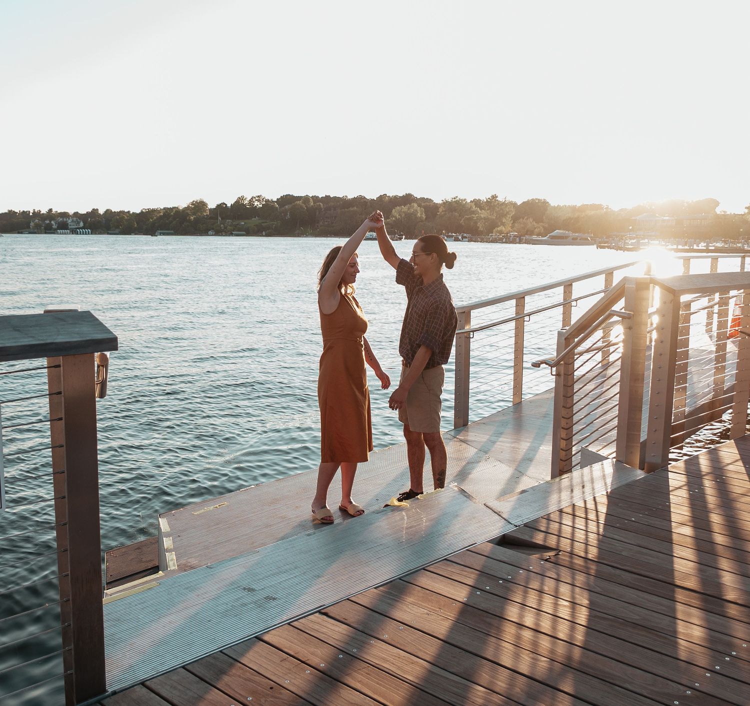 Couple dancing on a wooden dock at sunset with calm water and trees in the background.