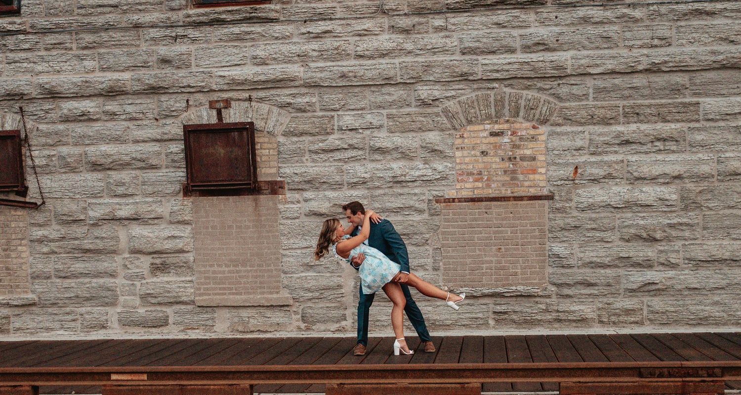 Romantic couple sharing a passionate dip kiss on a wooden deck against a rustic stone wall backdrop.
