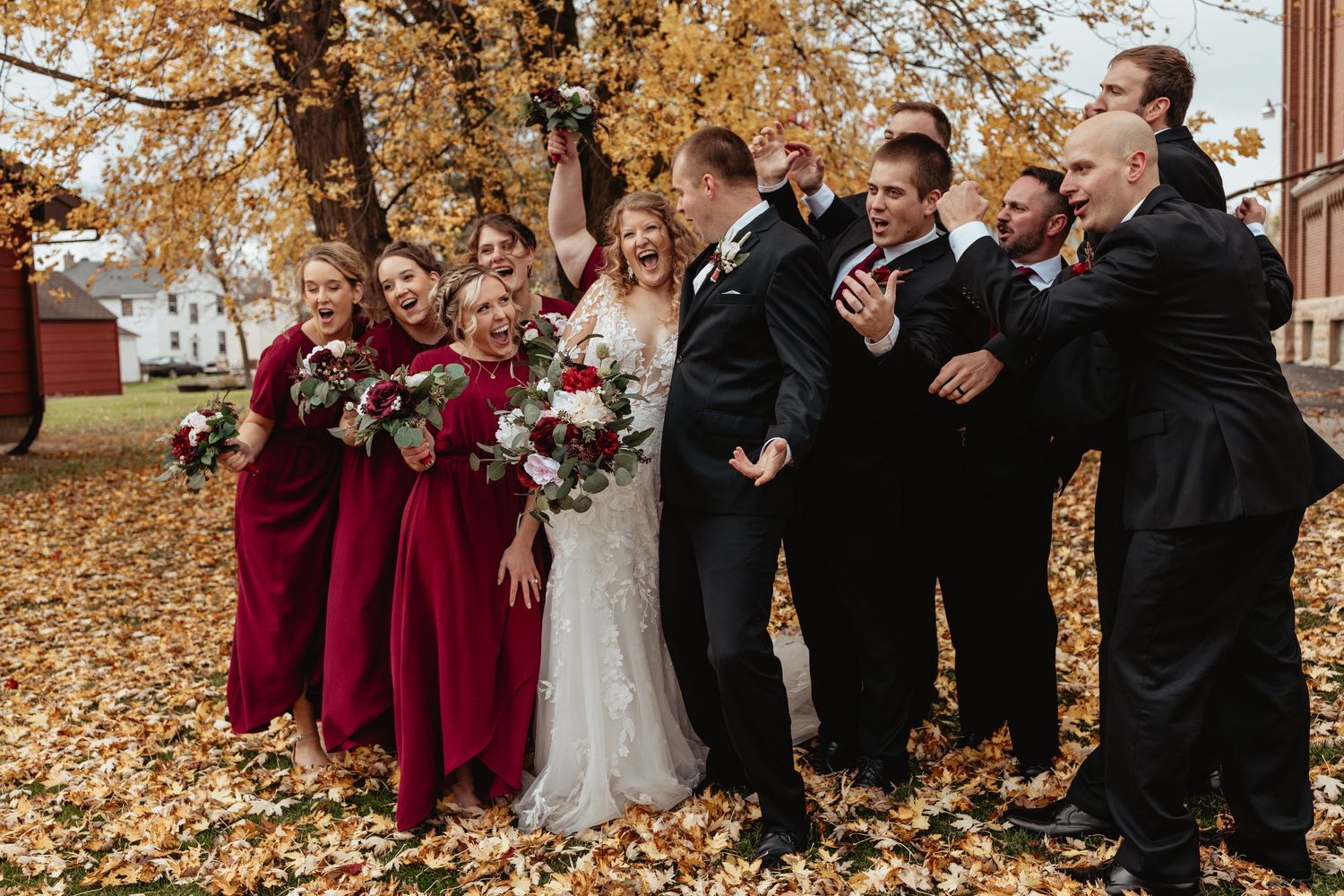 Wedding party poses together on fallen autumn leaves wearing burgundy and black formal attire.