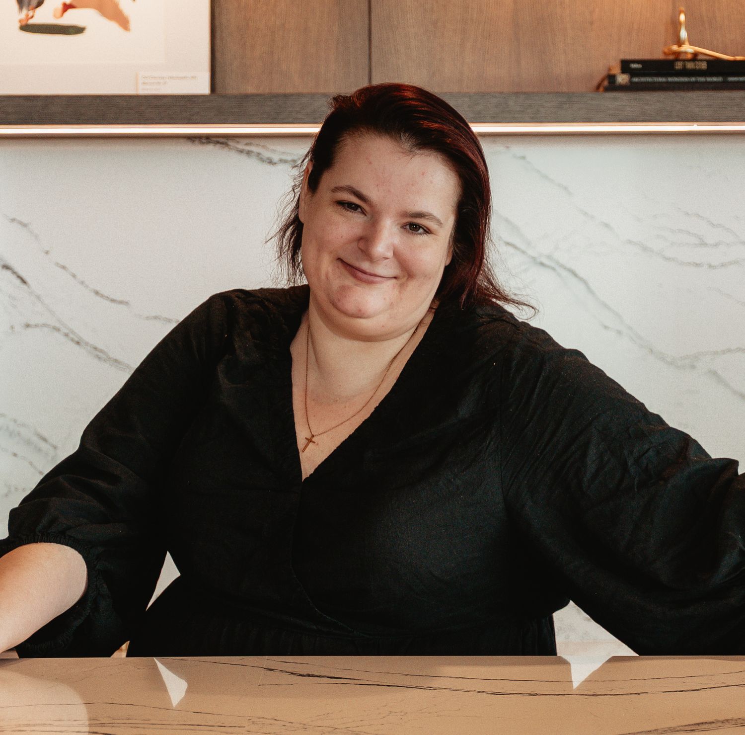 Professional in a black top seated at a desk with marble wall background, smiling warmly at the camera.