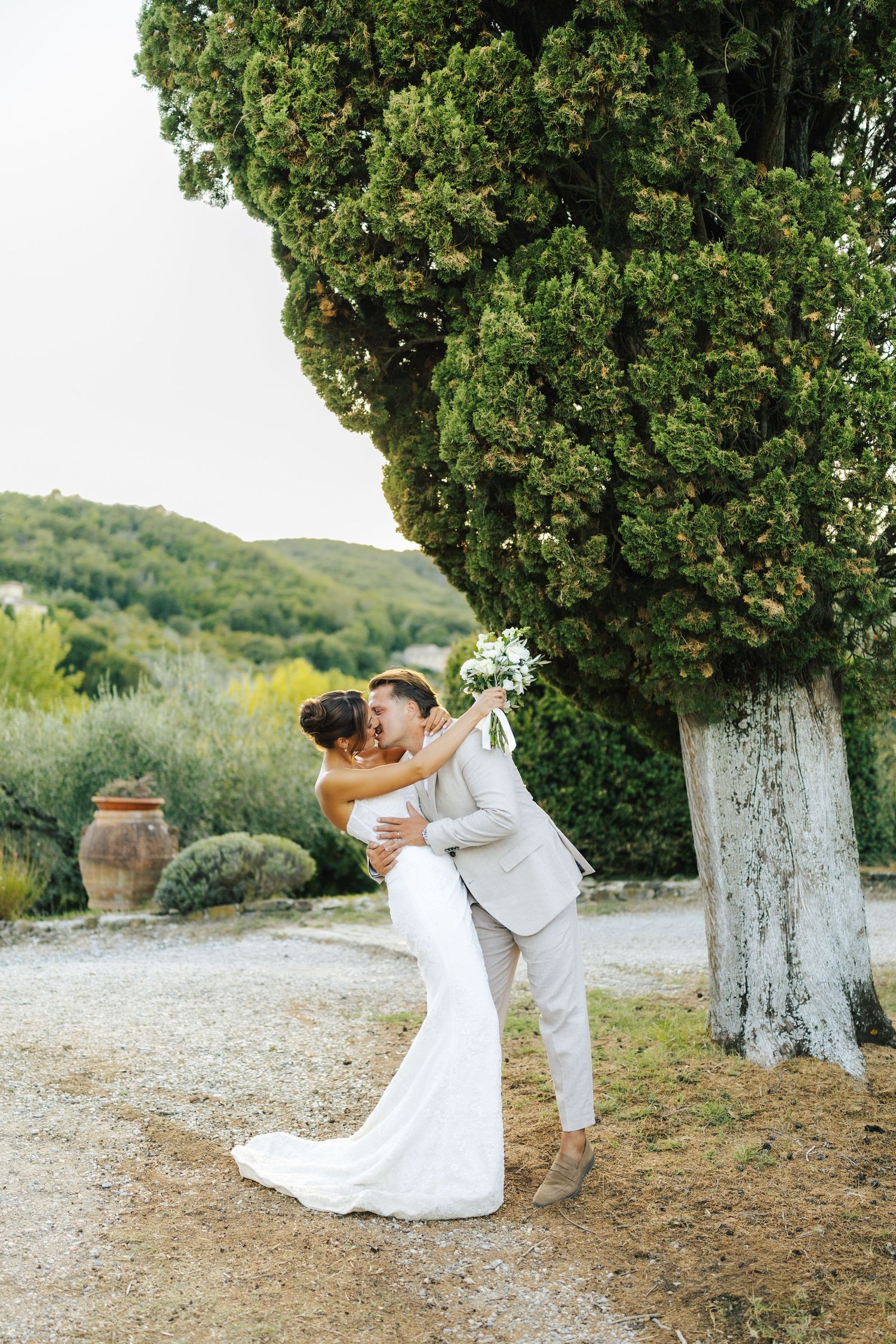 Nevěsta a ženich se ve svatební den romanticky objímají u velkého stromu v bujné toskánské zahradě.