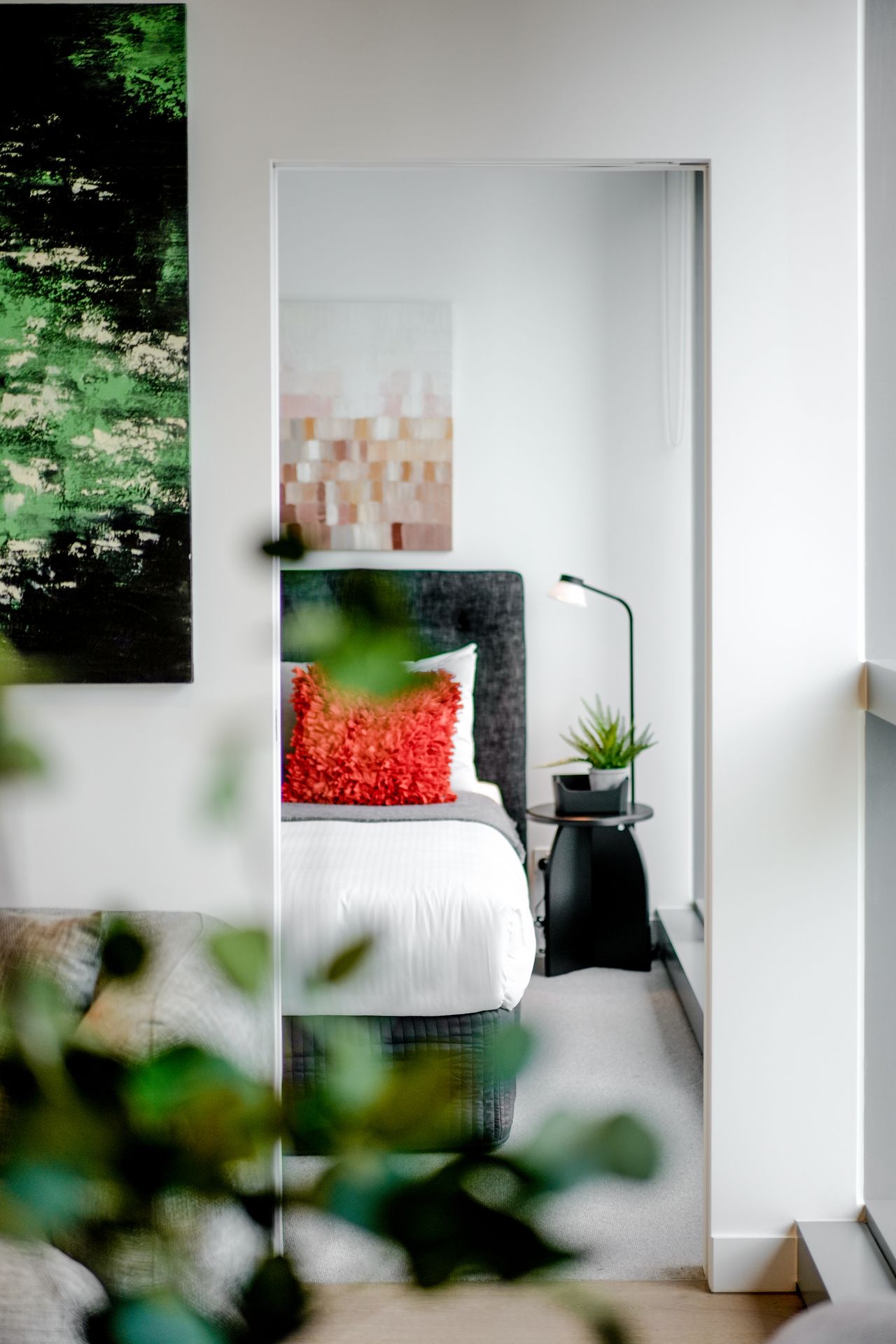 Minimalist bedroom interior with abstract wall art and plants visible through doorway.