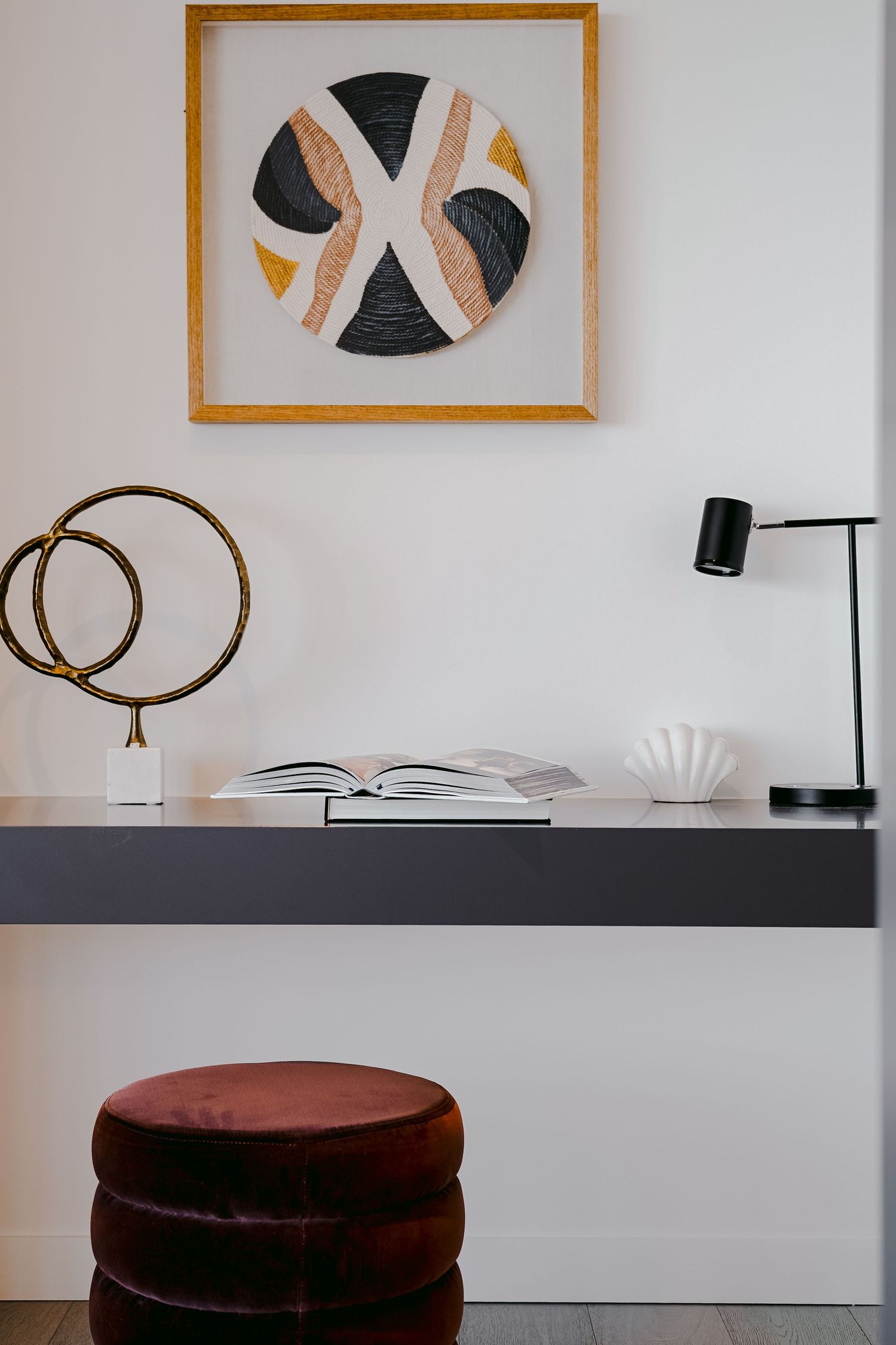 Minimalist desk setup with geometric framed artwork, task lamp, and burgundy velvet ottoman.