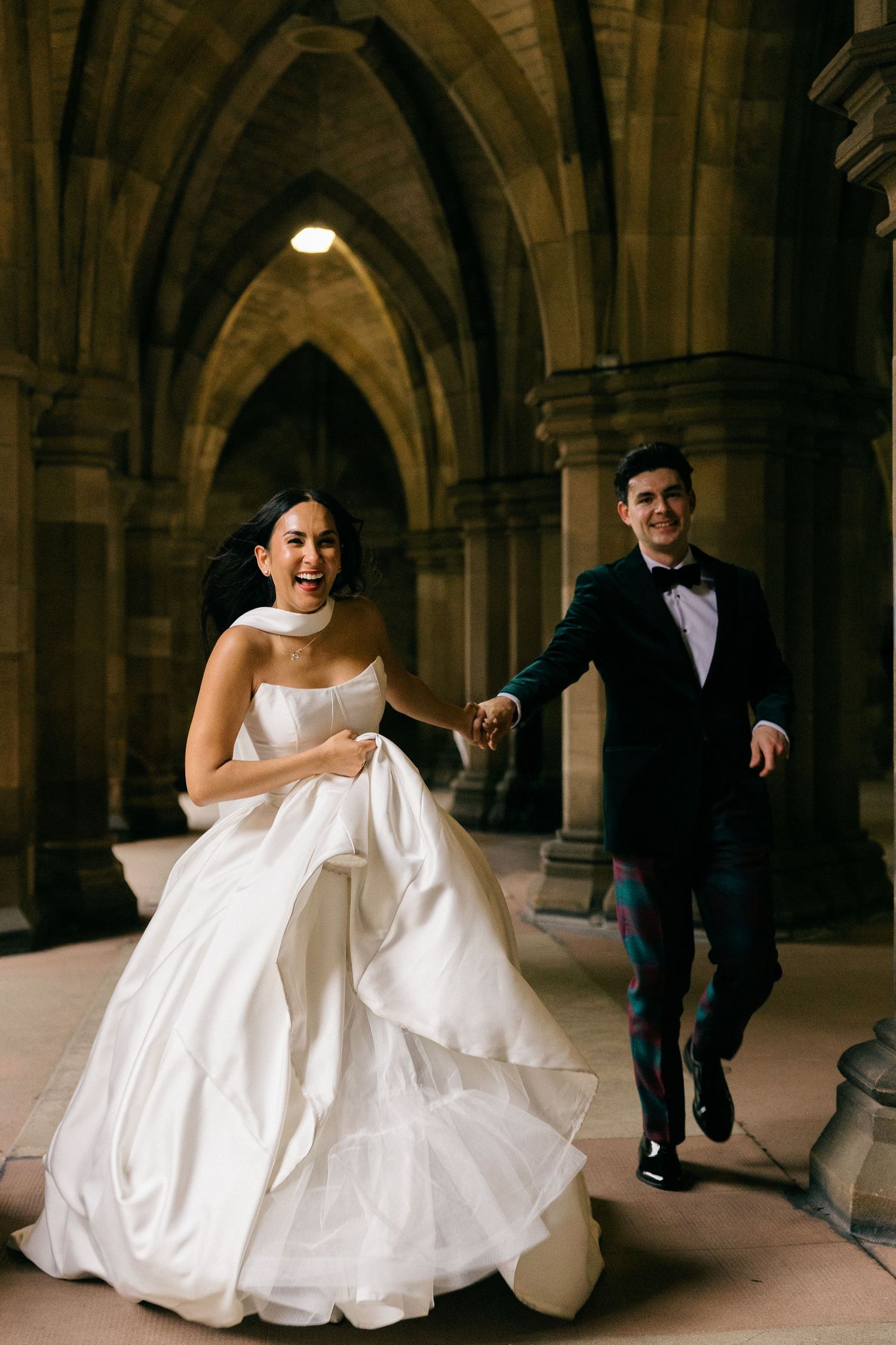 A joyful bride in a white gown and groom in a tuxedo run hand-in-hand through a stone archway.