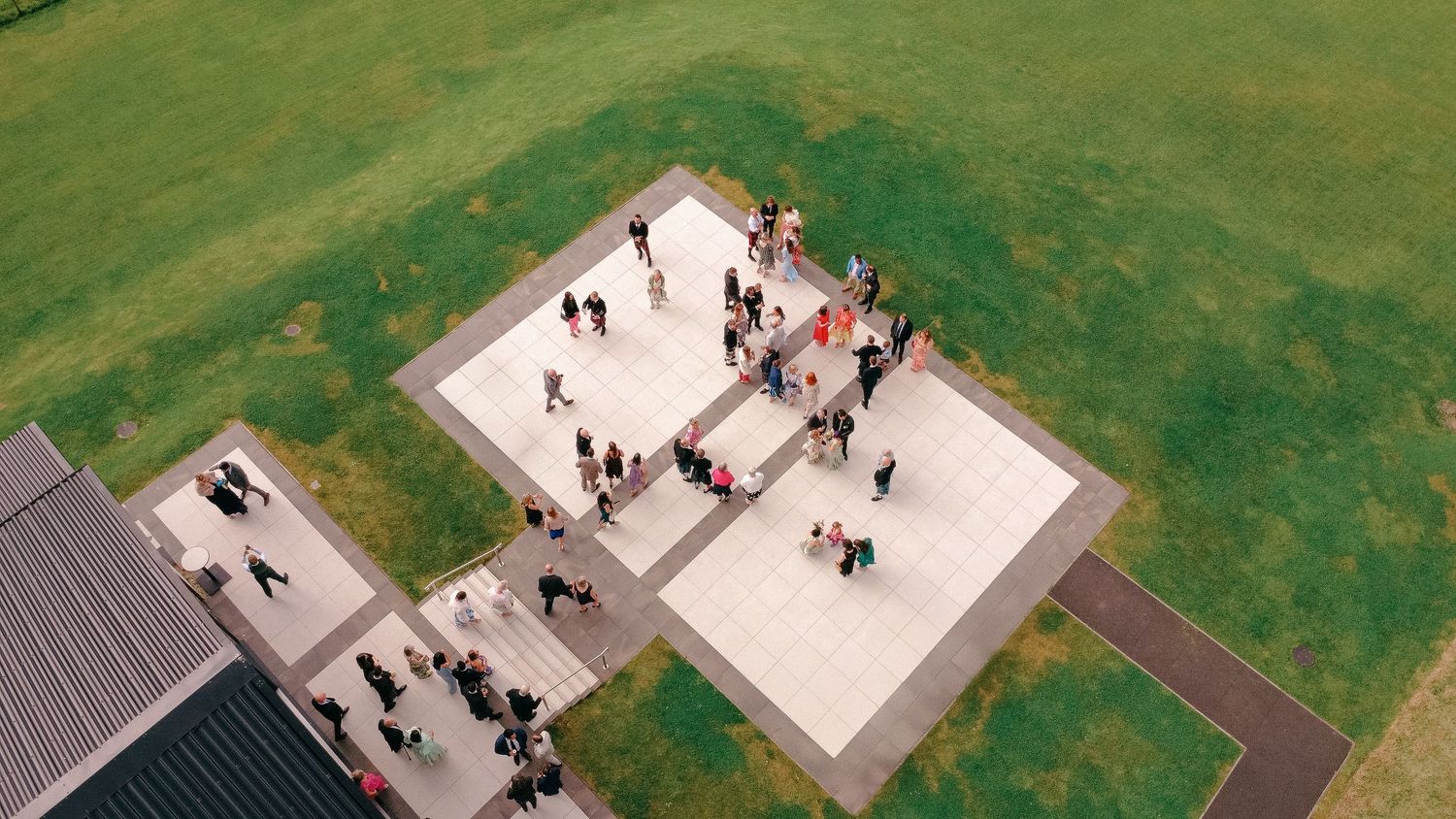 Aerial view of people gathering on a modern concrete plaza surrounded by green grass near a building.