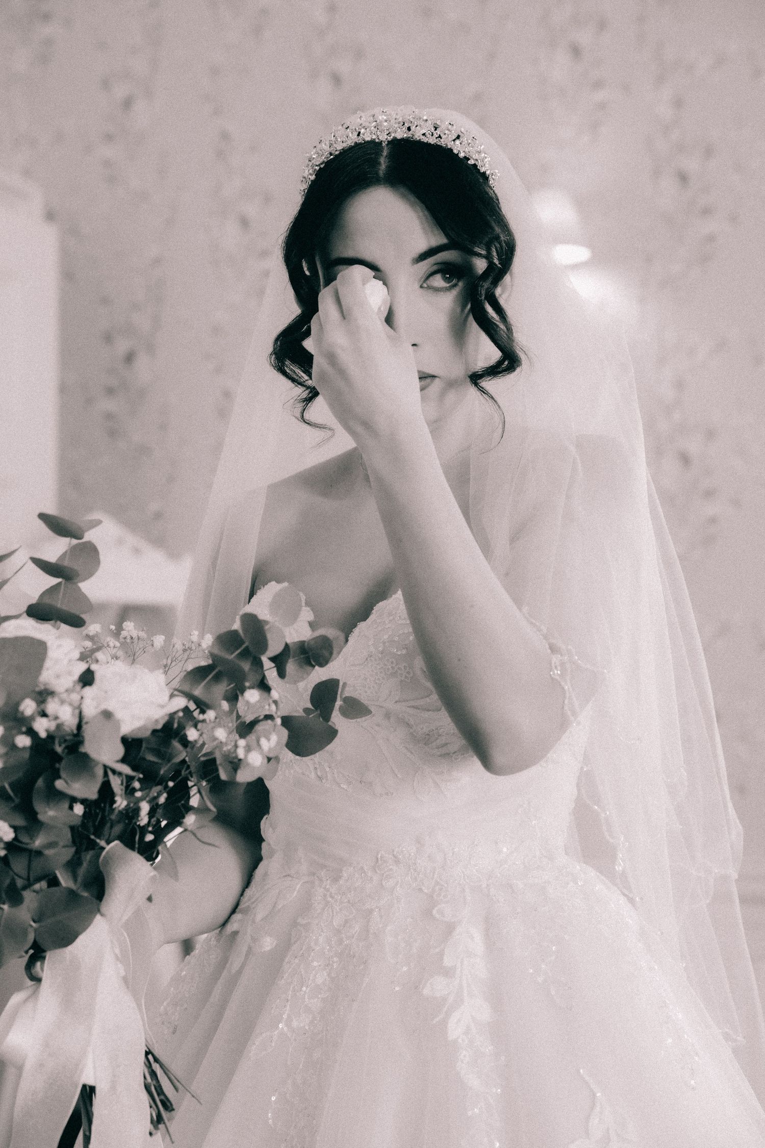 A bride in a white lace wedding gown and veil holds a bouquet while covering part of her face in black and white.