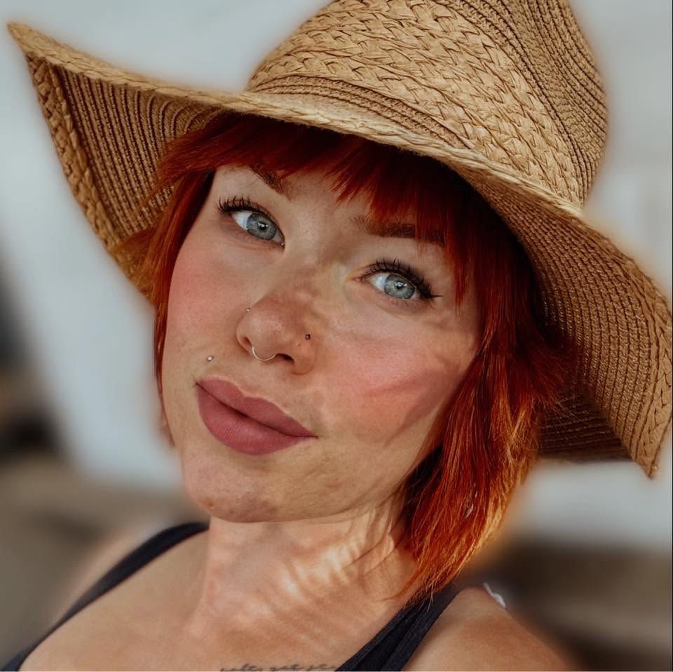 A headshot of a slightly edge and beautiful red-headed woman wearing a straw hat.