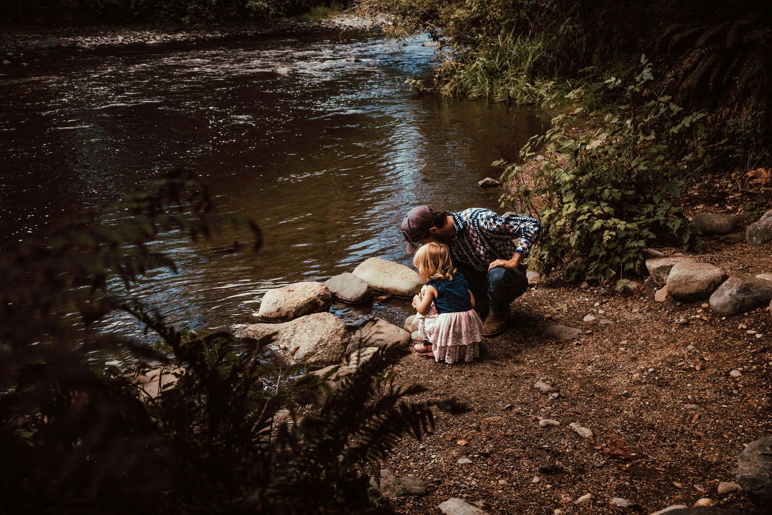 A man pointing something out to his daughter, as they crouch by the Quinsam River.