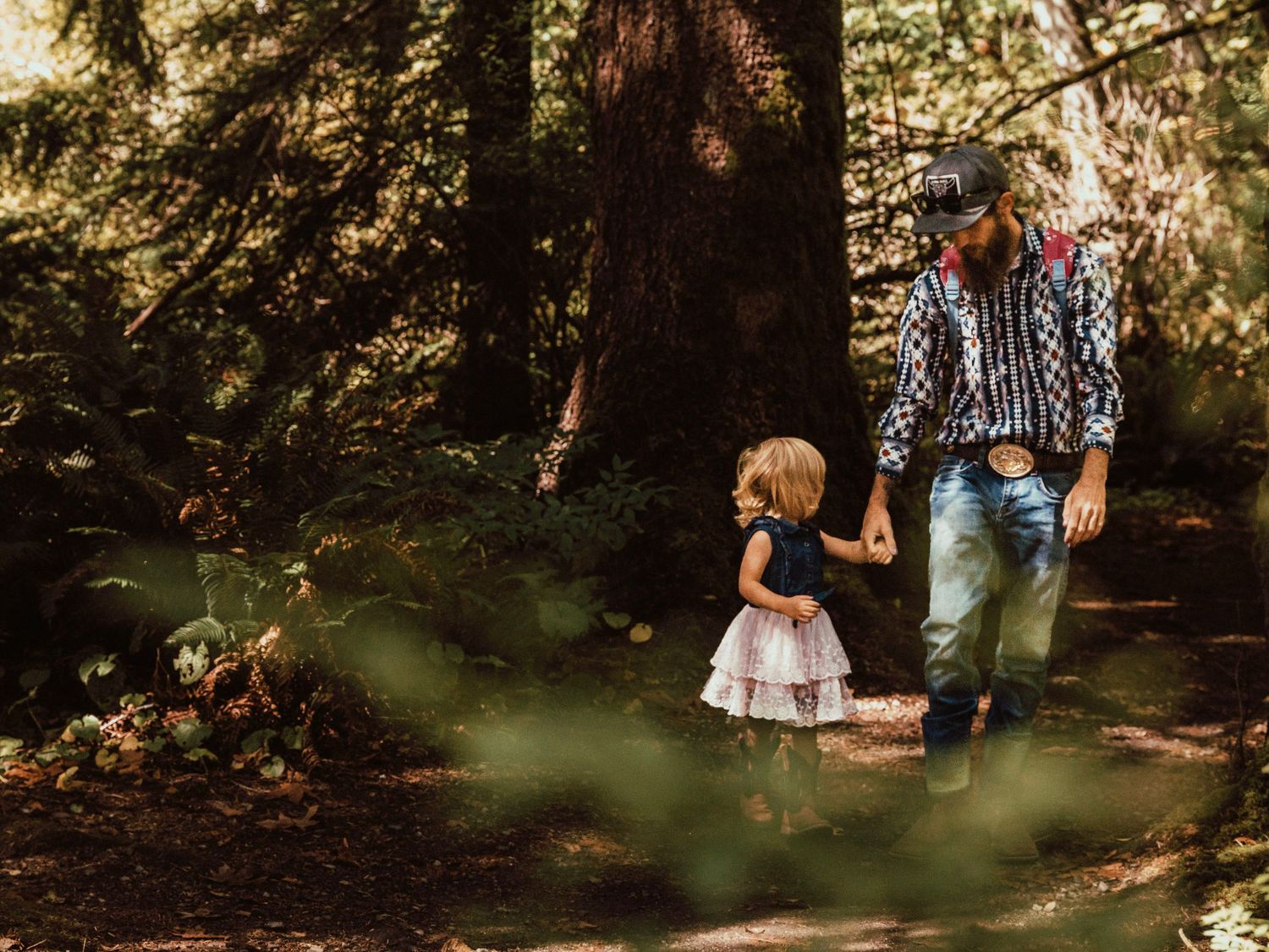 A cowboy and his daughter walking holding hands through a forest.