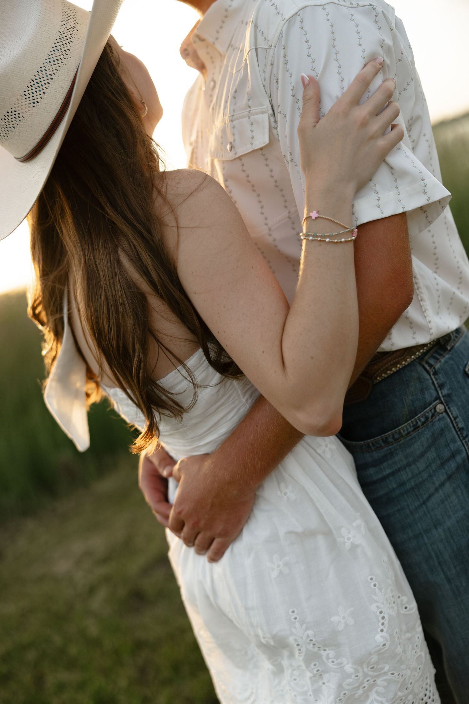 Bride in white lace dress wearing cowboy hat embracing groom in jeans at golden hour sunset.