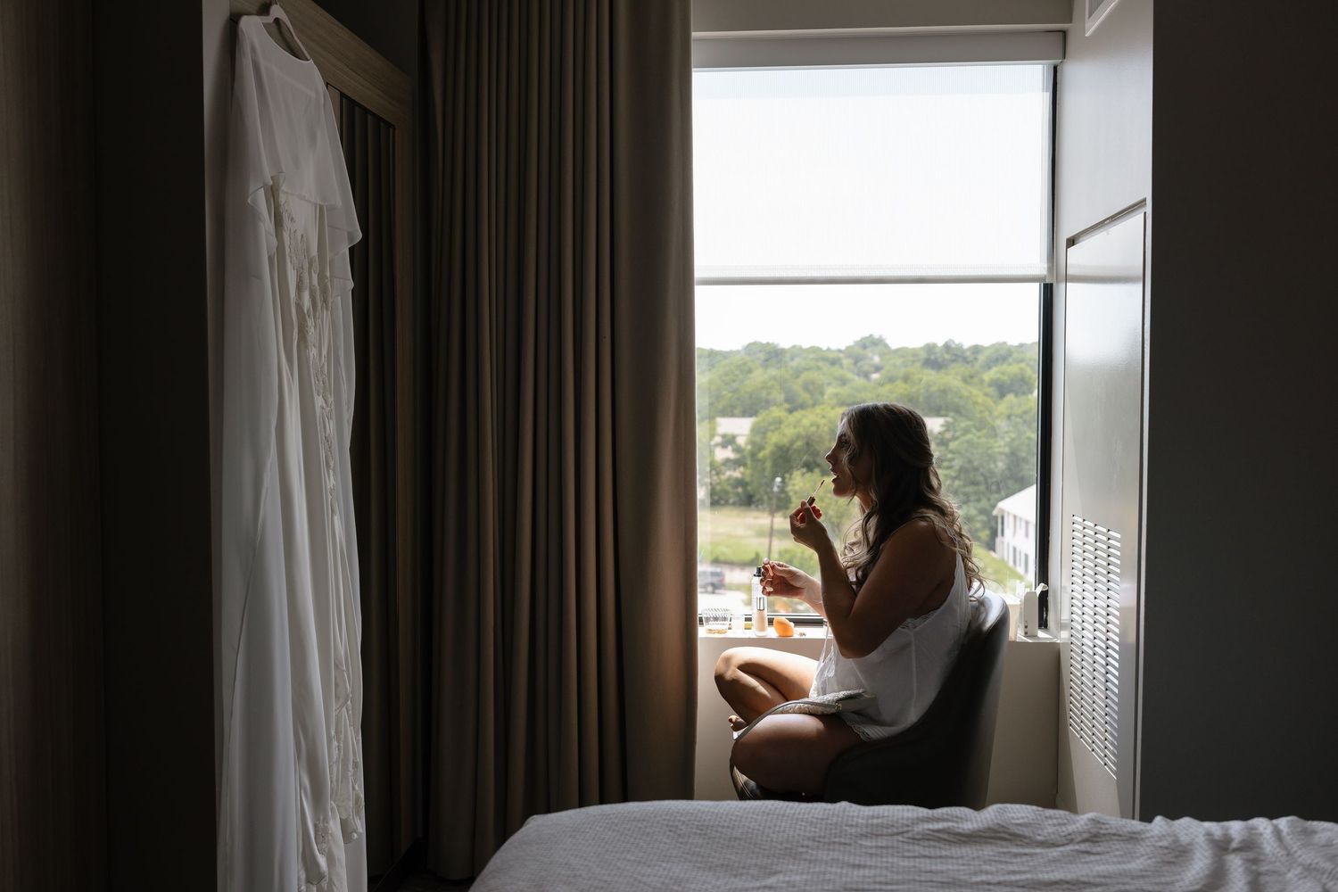 Bride sits by hotel window sipping drink beside hanging wedding dress before ceremony.