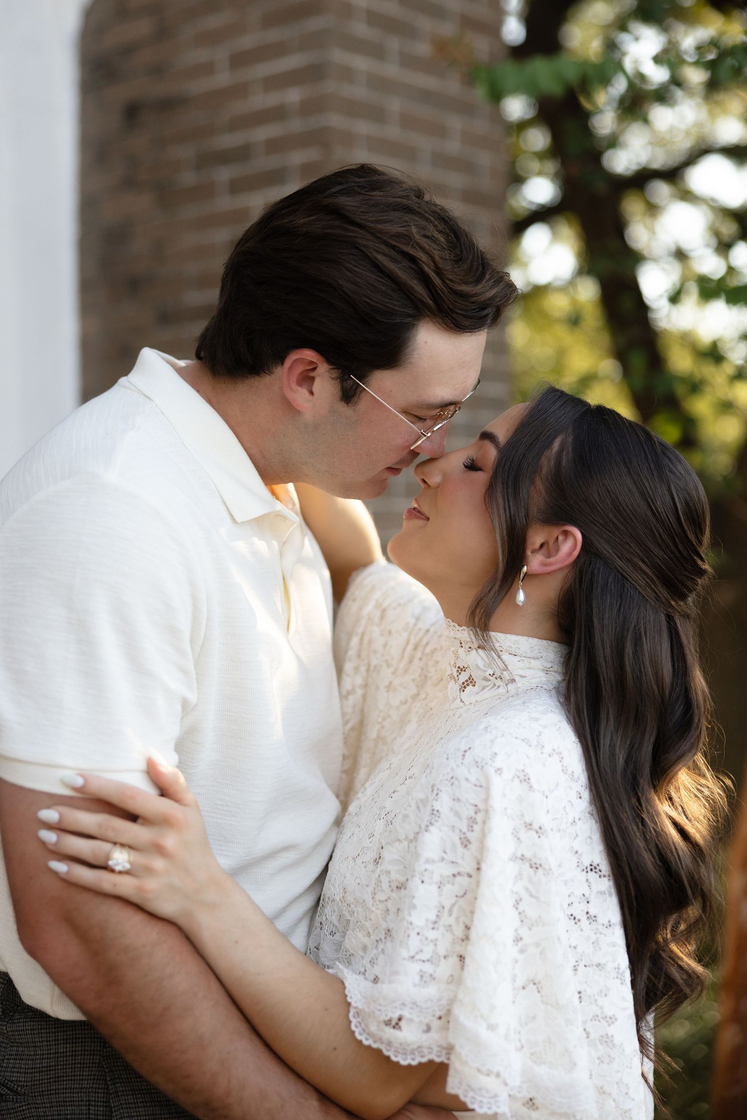 A couple shares a kiss outdoors, the woman wearing a white lace dress and sparkling engagement ring.