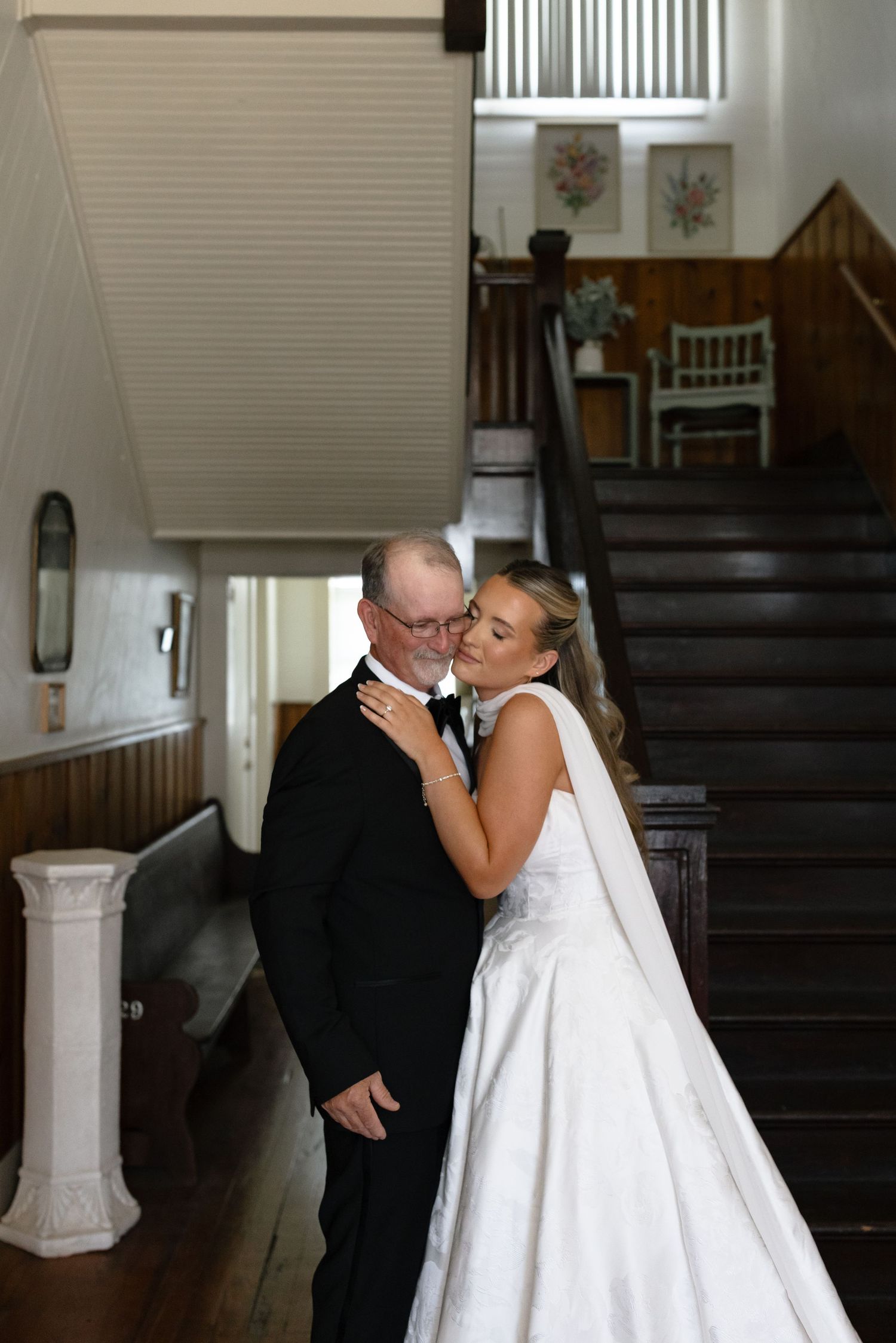 Bride in white ball gown kissing older gentleman in tuxedo near elegant wooden staircase indoors.