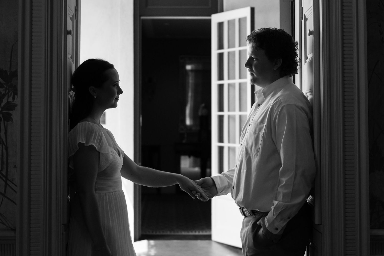 Black and white photo of a bride and groom sharing a tender handshake moment in a doorway.