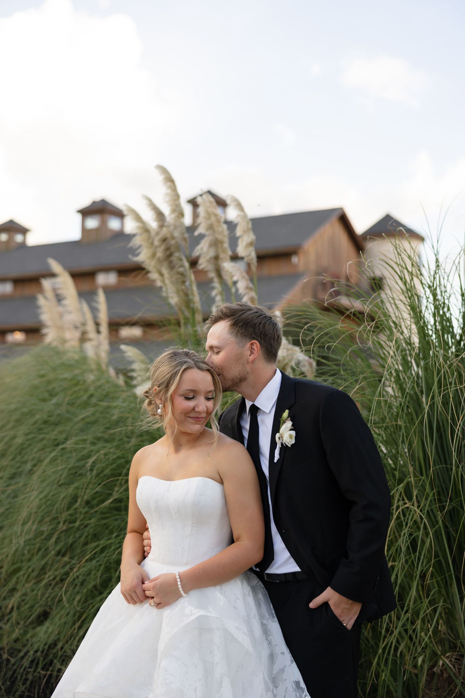 Groom in black tuxedo kissing bride on cheek outdoors near tall pampas grass at rustic wedding venue.