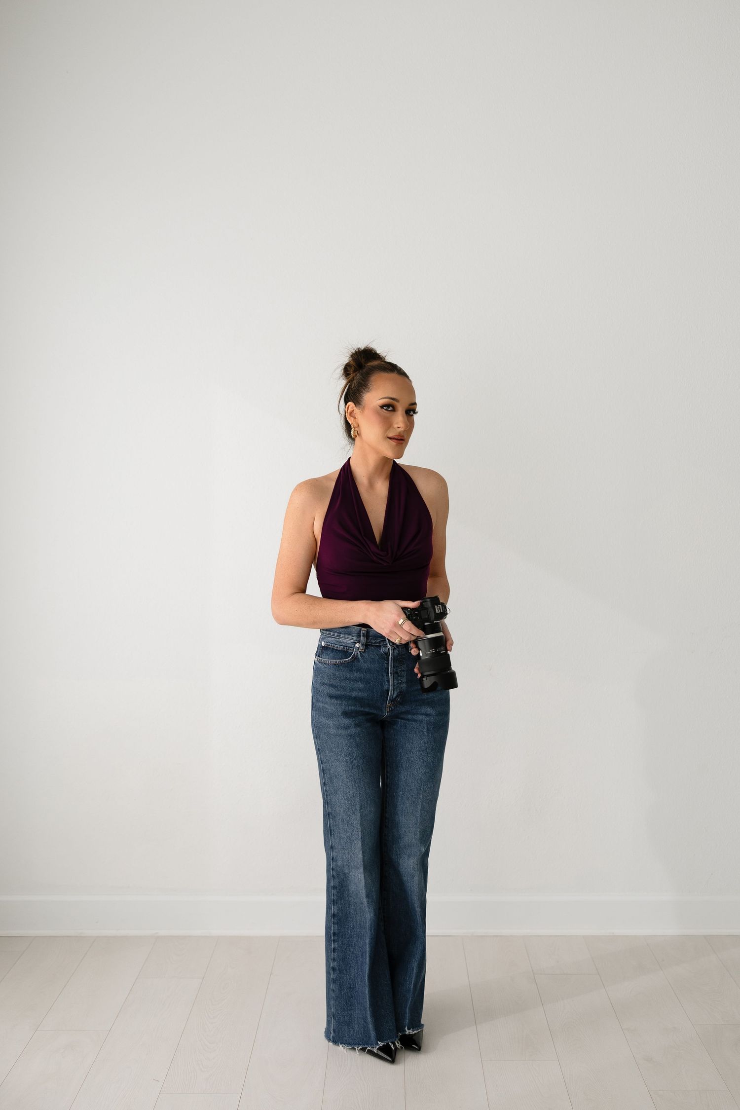 Female photographer in burgundy halter top and jeans holding camera against a clean white studio wall.