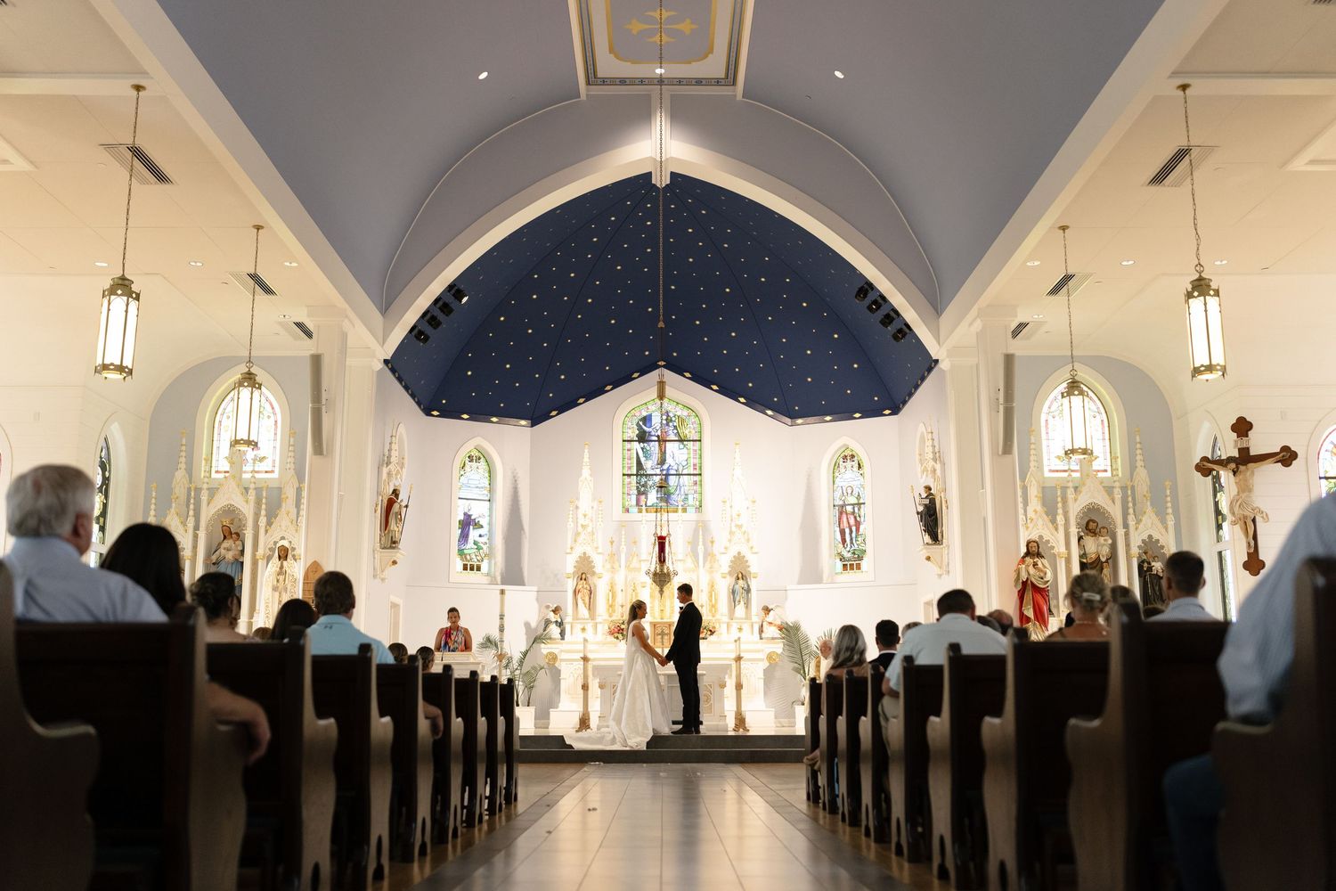 Wedding ceremony inside white church with blue vaulted ceiling, stained glass windows, and guests in wooden pews.