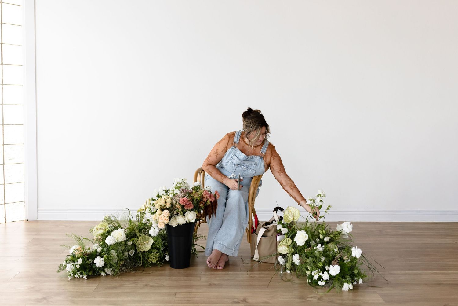 Florist arranging elegant white and blush wedding floral installation on wooden floor against white wall.