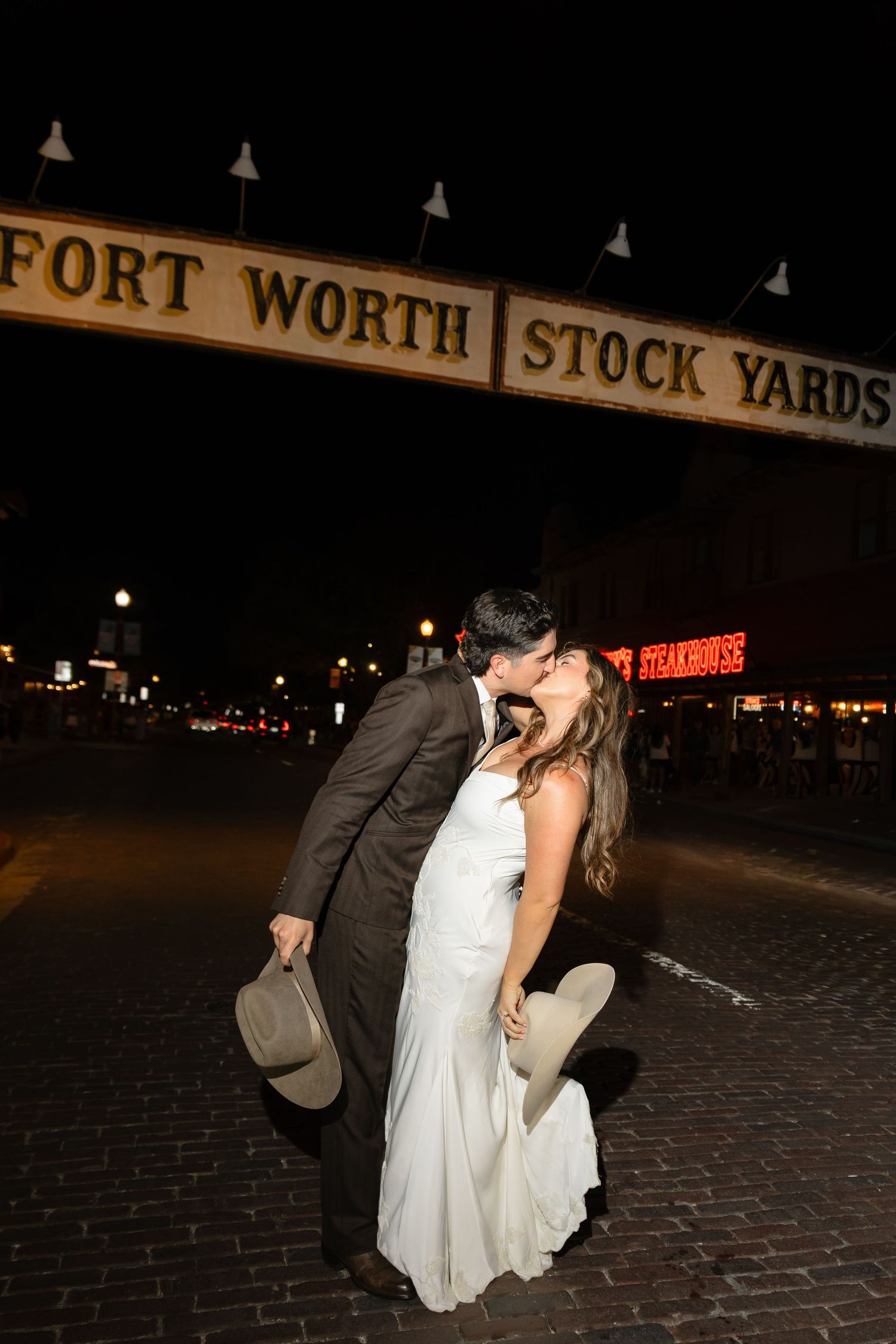 Bride and groom sharing romantic dip kiss under Fort Worth Stockyards sign at night.