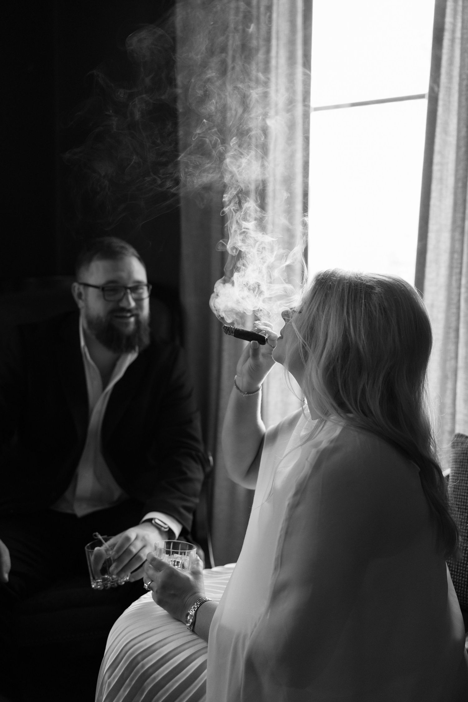 Black and white photo of a bride smoking a cigar and holding a drink while a bearded groom smiles beside her.