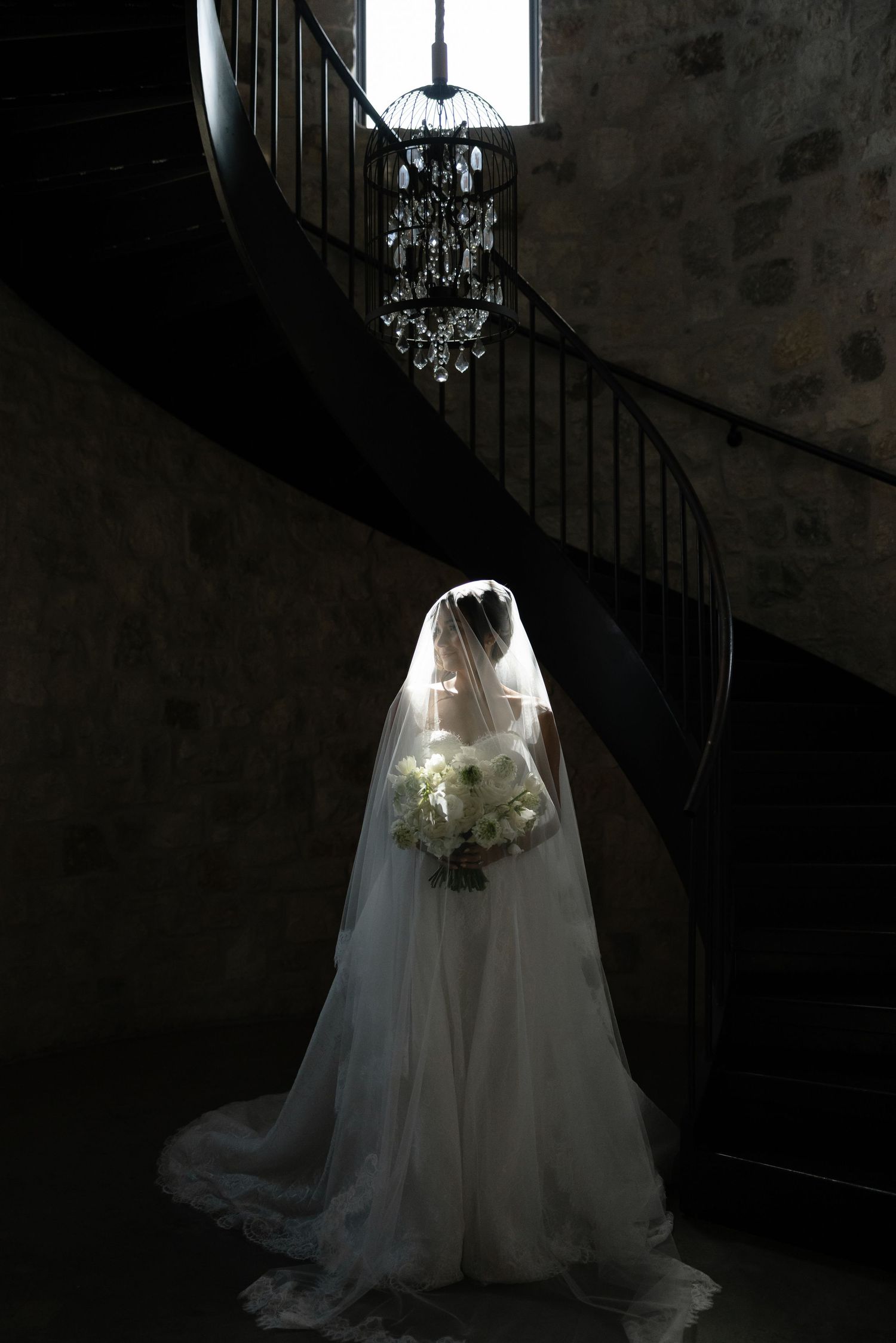 Bride in white gown and veil holds bouquet beneath crystal chandelier at base of dark spiral staircase.