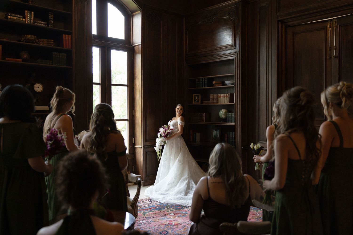 Bride in white gown stands before tall windows as bridesmaids look on in dark library.