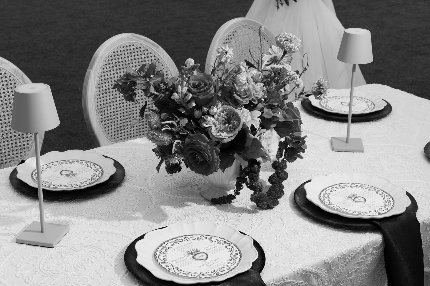 Elegant outdoor wedding table setting with floral centerpiece, decorative plates, and small table lamps in black and white.