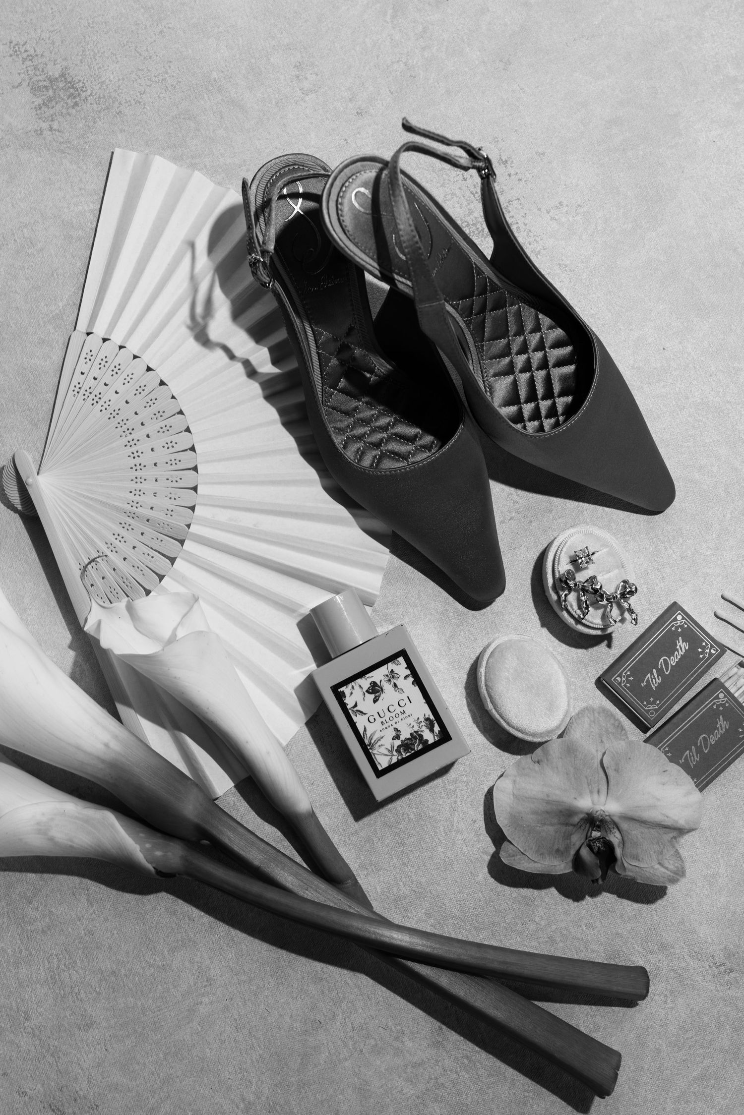 Black and white flatlay of Gucci perfume, heels, hand fan, orchid, and accessories on concrete.