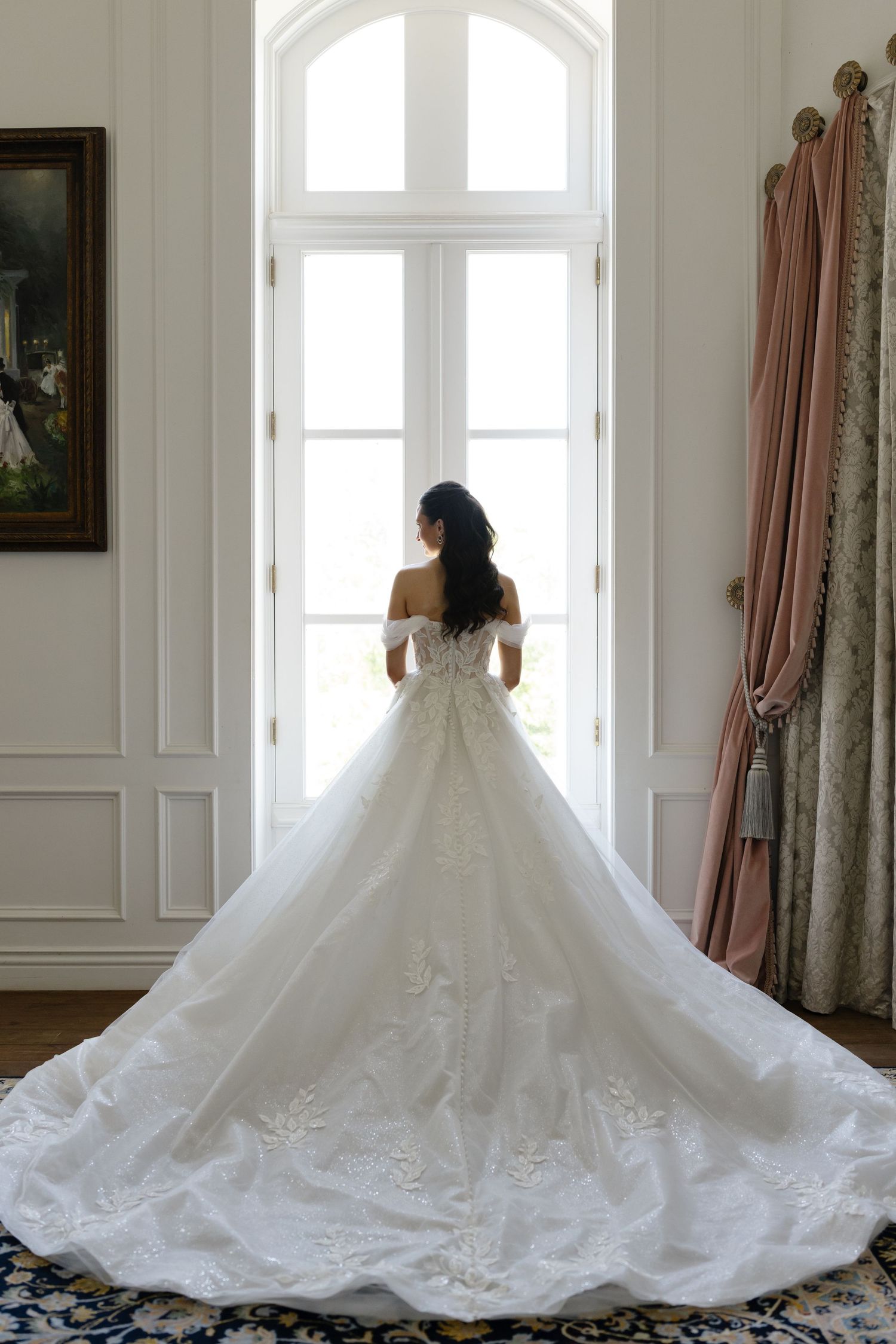 Bride in elegant off-shoulder ball gown with long lace train stands before tall window in ornate room.