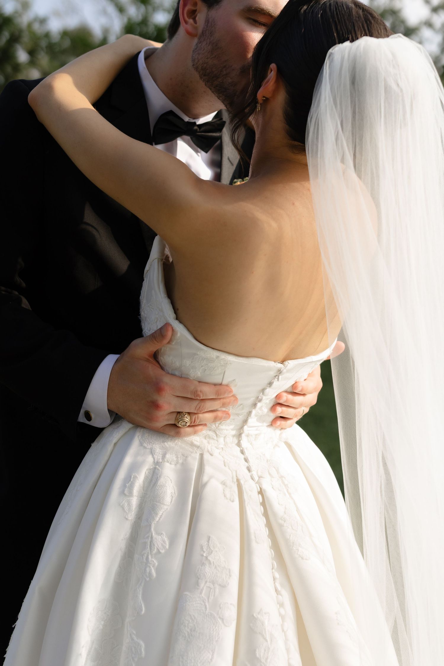 Groom in black tuxedo embracing bride in backless lace wedding gown with veil outdoors.