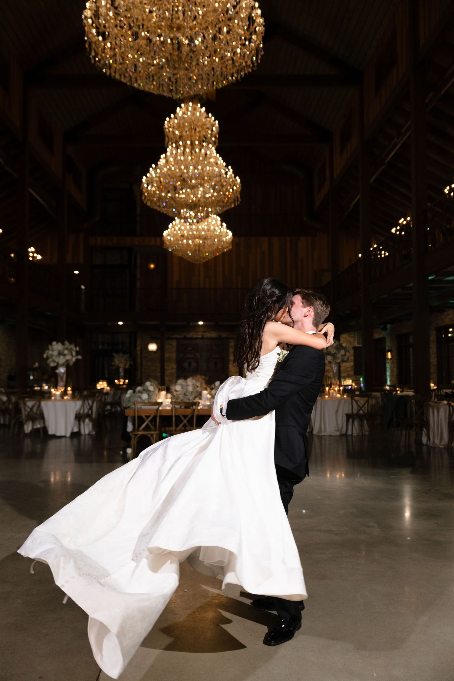 A groom lifts his bride during their first dance under grand crystal chandeliers in a rustic barn venue.