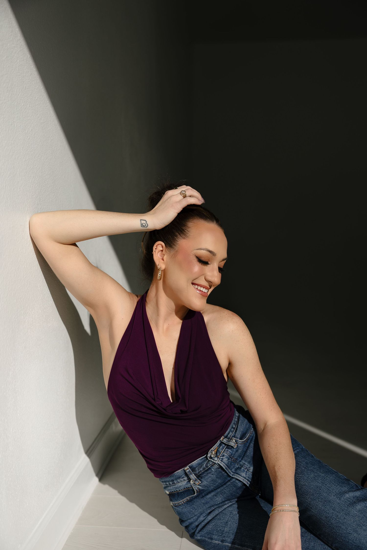 Smiling woman in purple halter top and jeans leaning against white wall with hand raised to head.