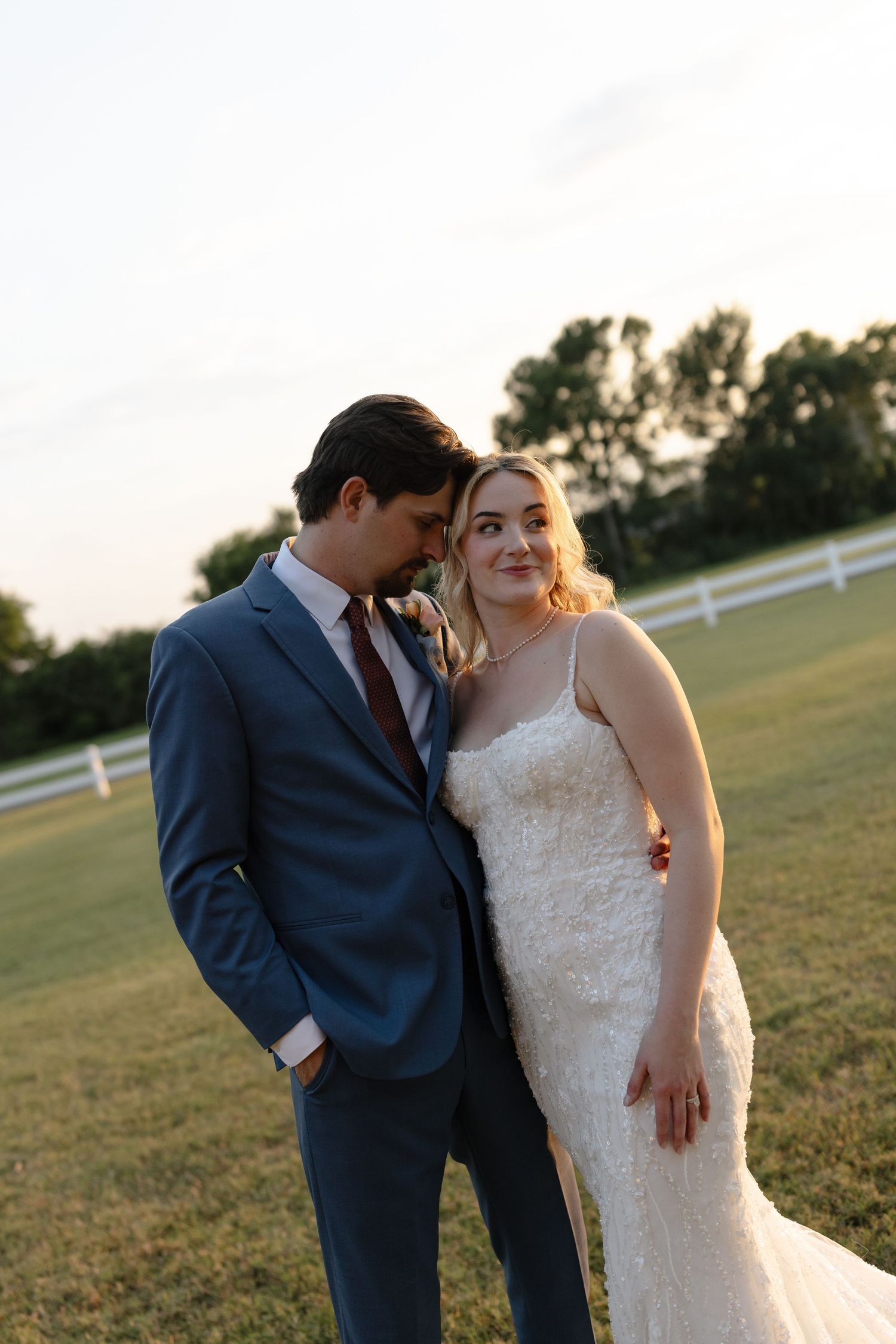 A bride in a lace gown and groom in a blue suit share a romantic moment at golden hour in a green field.