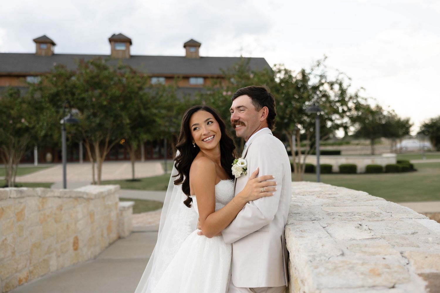 Bride and groom embracing outdoors at wedding venue, smiling couple in white dress and tan suit.