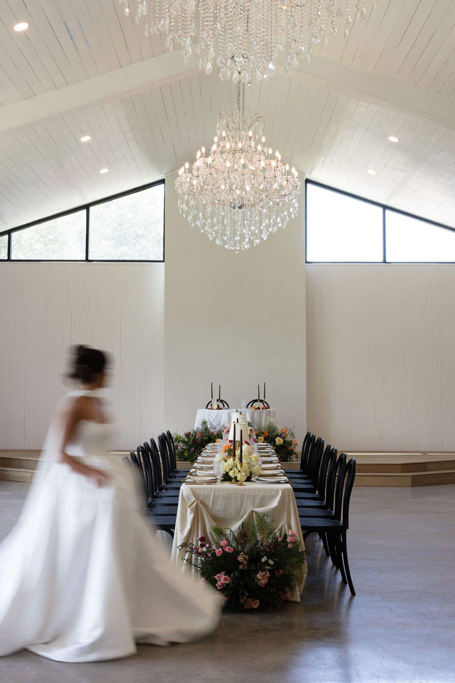 Bride in white gown walks past elegant chandelier-lit reception hall with floral centerpiece table.