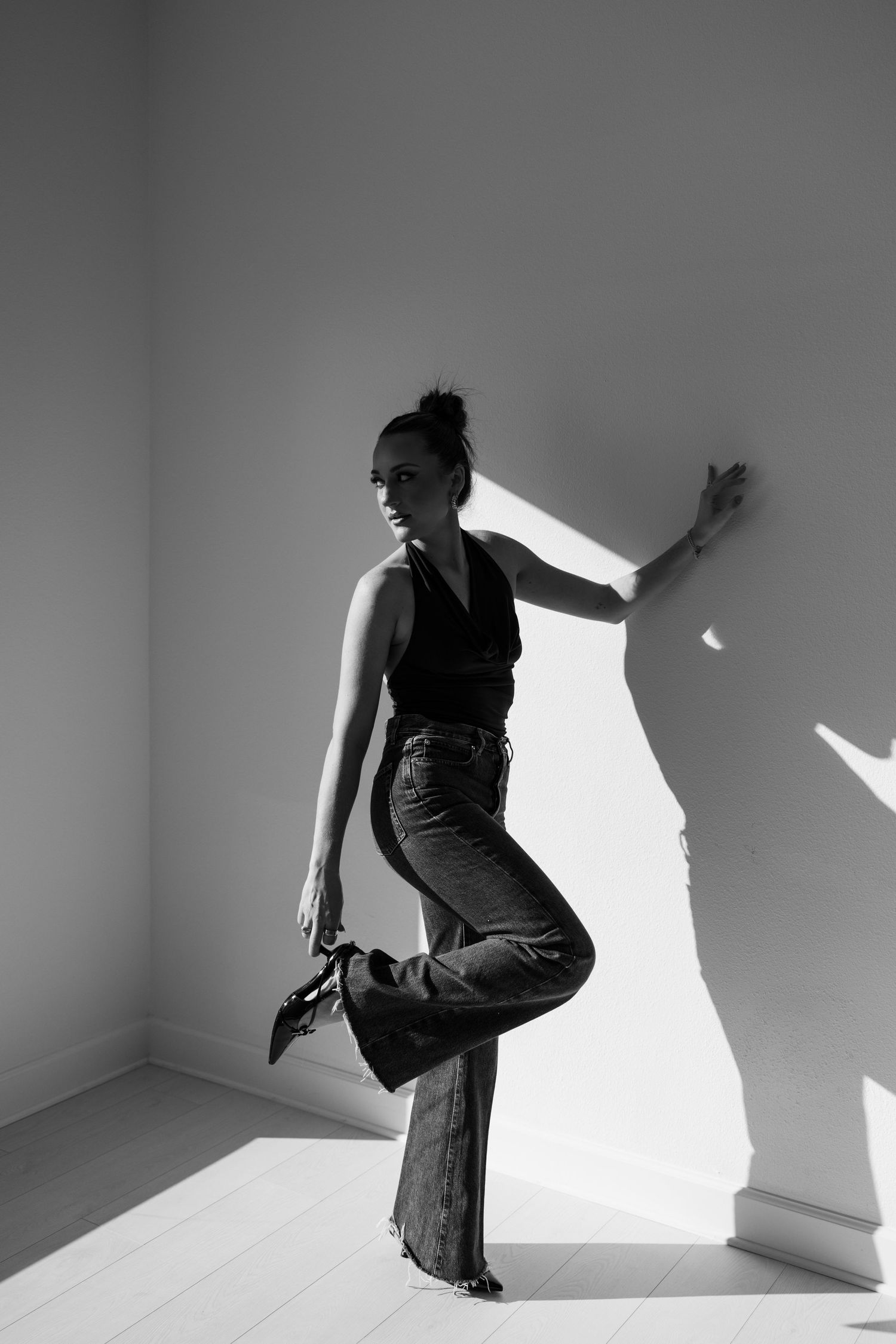 Black and white artistic portrait of woman in halter top and jeans posing against sunlit white wall.
