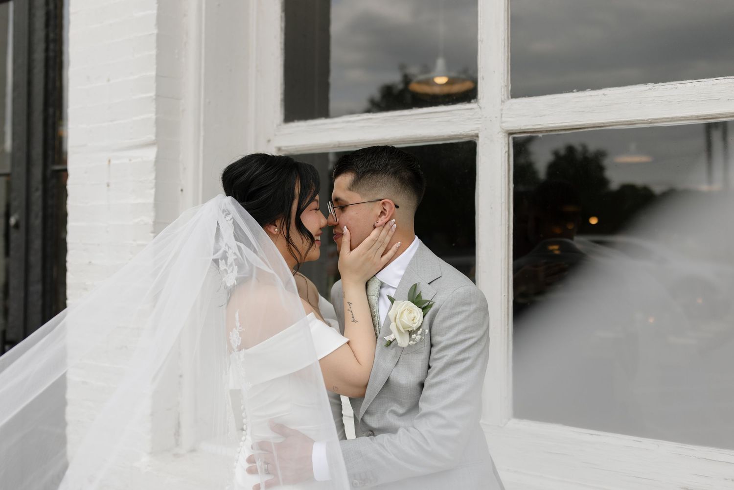 Bride and groom share a romantic kiss beside a white window on their wedding day.