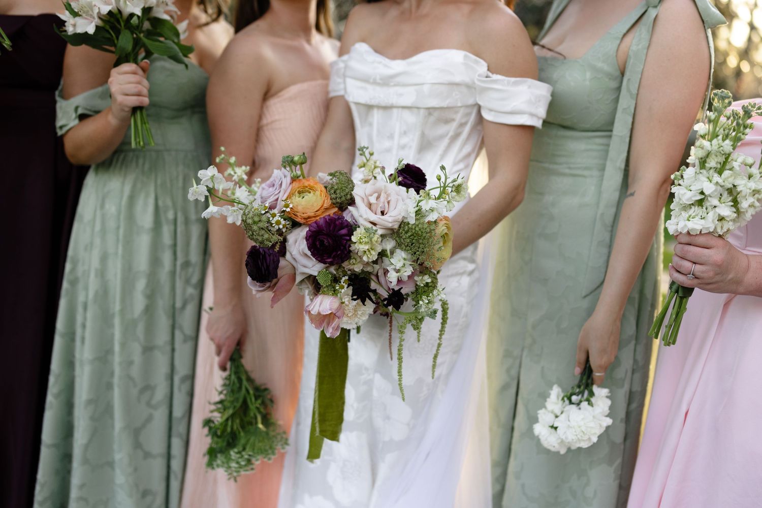 Bride in white off-shoulder gown with bridesmaids in sage and blush dresses holding floral bouquets.