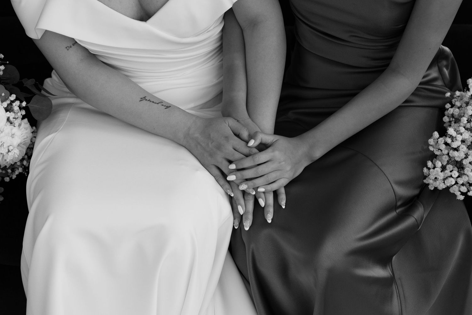 Black and white close-up of bride and bridesmaid sitting together holding hands in white and gray gowns.
