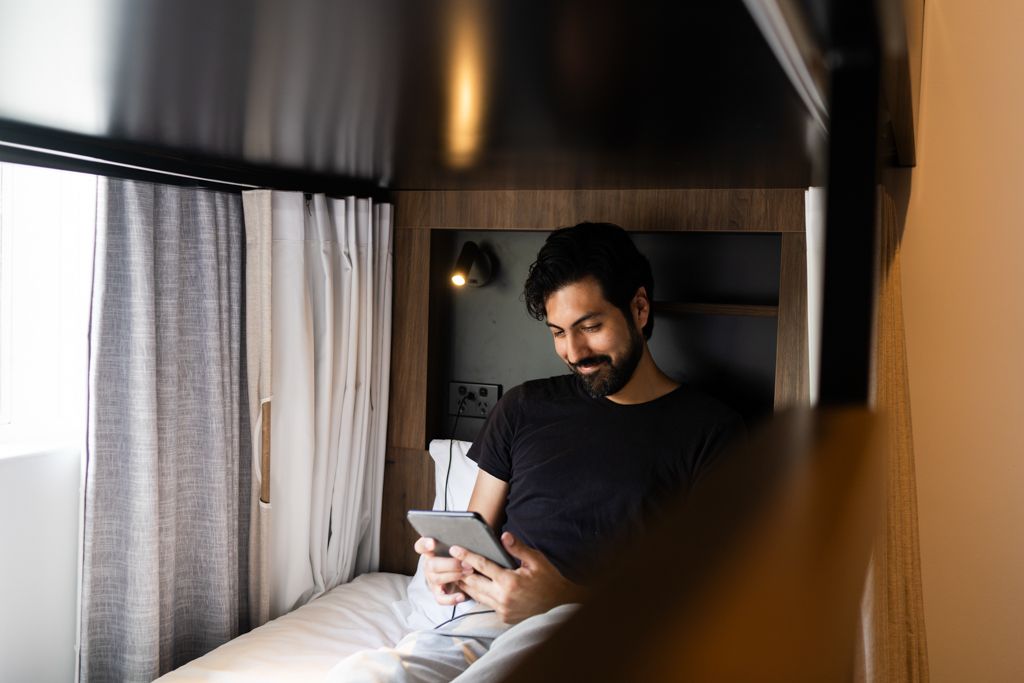 Bearded man relaxing on bed in small cabin using a tablet device.