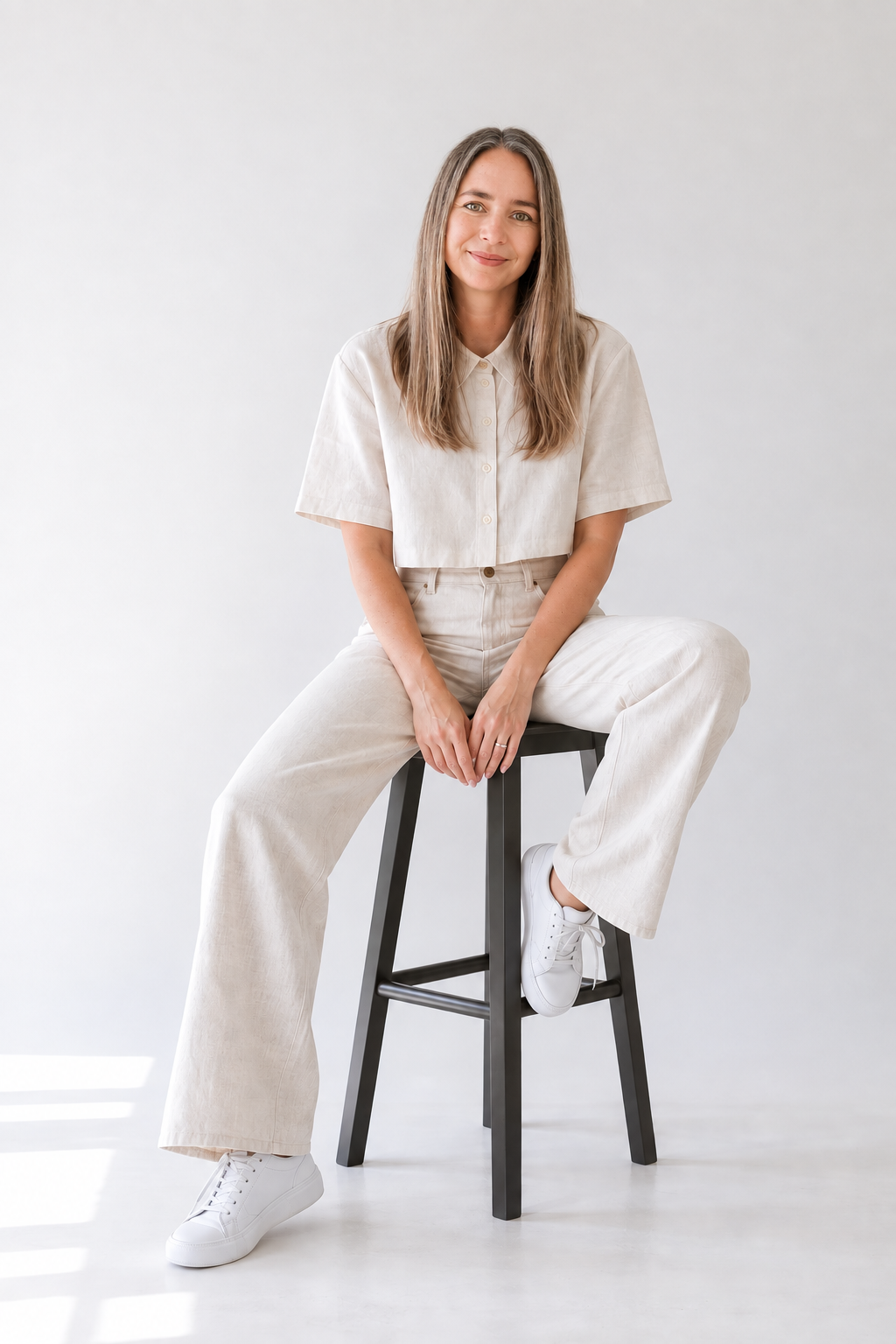 Smiling woman in beige short-sleeve top and wide-leg pants seated on a black stool against white background.