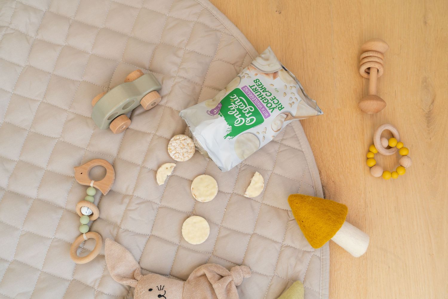 Baby play mat with wooden toys, teethers, a mushroom rattle, and organic snacks arranged on a quilted beige surface.