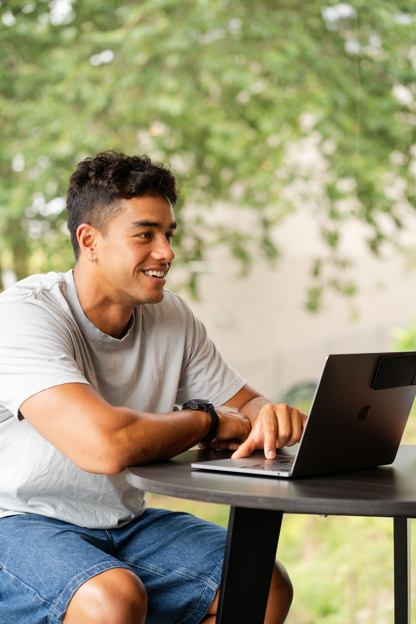 Smiling young man working on laptop at outdoor table surrounded by lush green trees.