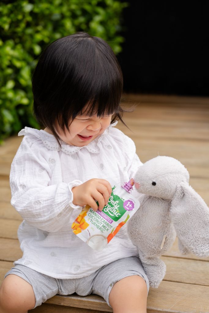 Smiling Asian toddler in white blouse holding an Only Organic food pouch with a stuffed bunny.